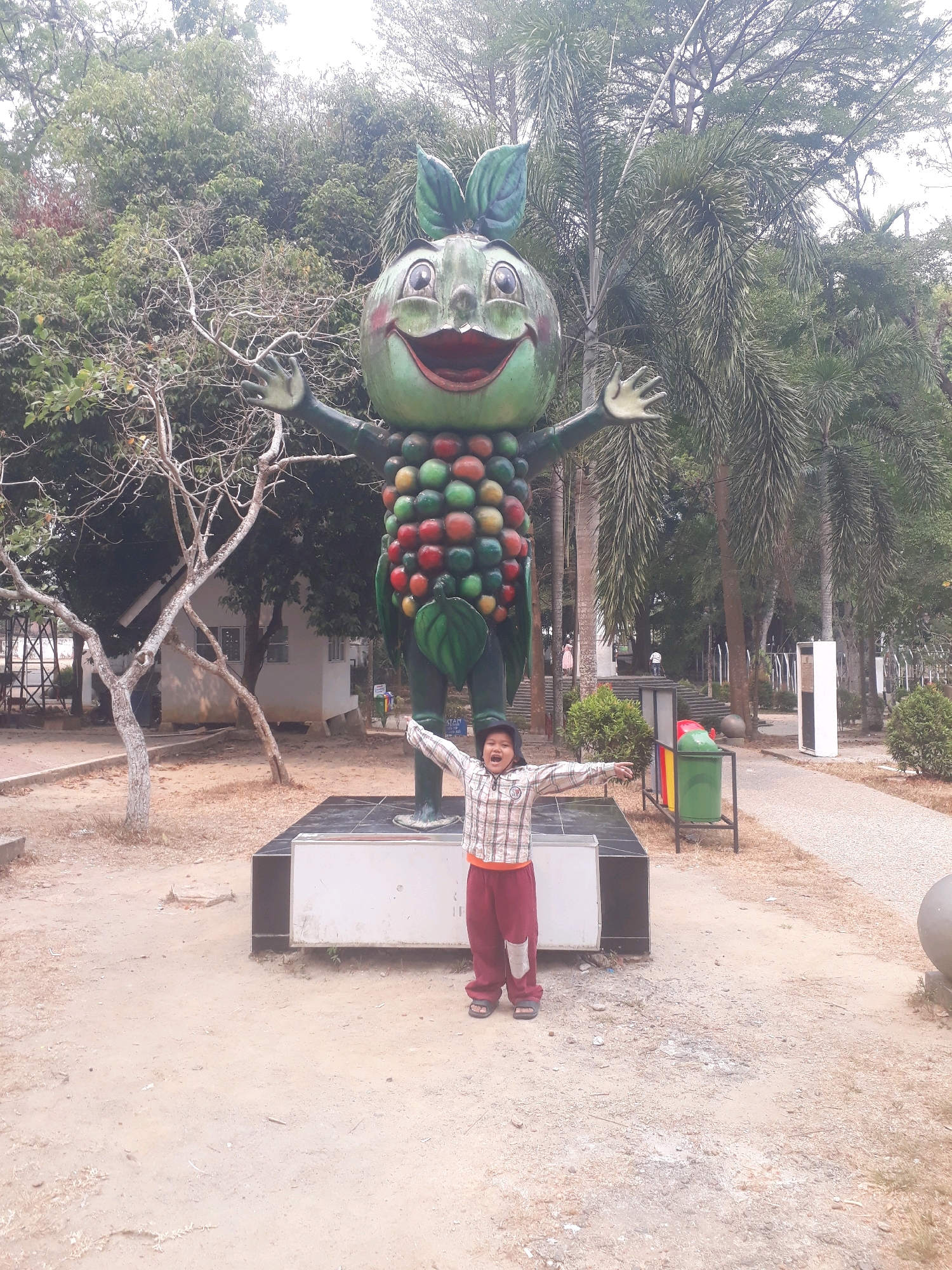 A child stands with arms outstretched in front of a large, cartoonish statue of a green fruit resembling a pomegranate, with a smiling face and hands raised. The statue is adorned with numerous colorful, round fruits, and stands on a black and white base. The child is wearing a plaid shirt, maroon pants with a patch, and sandals. The scene is set in an outdoor park with trees, some of which are palm trees. In the background, there are buildings, a trash receptacle with multiple compartments, and a gravel pathway. The ground is covered with sand and dry leaves. The image appears to be taken during the day, with overcast lighting. The statue's text is illegible. The location of the statue is not specified.