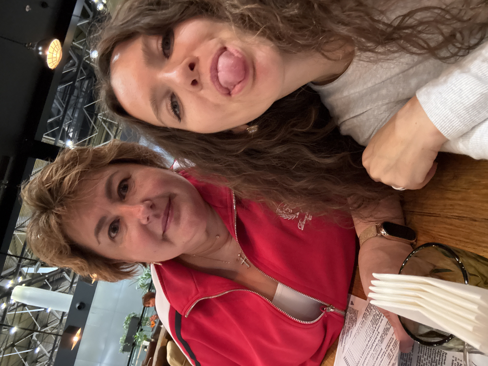 Two women are taking a selfie at a table in a restaurant. The woman on the left is older, with short brown hair and is wearing a red jacket over a white top. She is wearing a cross necklace. The woman on the right is younger, with long curly brown hair. She is sticking her tongue out at the camera and is wearing a light gray sweater. In the foreground, there is a glass with napkins, a menu, and a watch on the left woman's wrist. The background shows a restaurant interior with exposed ceilings and lighting fixtures. The location is identified as a restaurant in Khimki, Russia.
