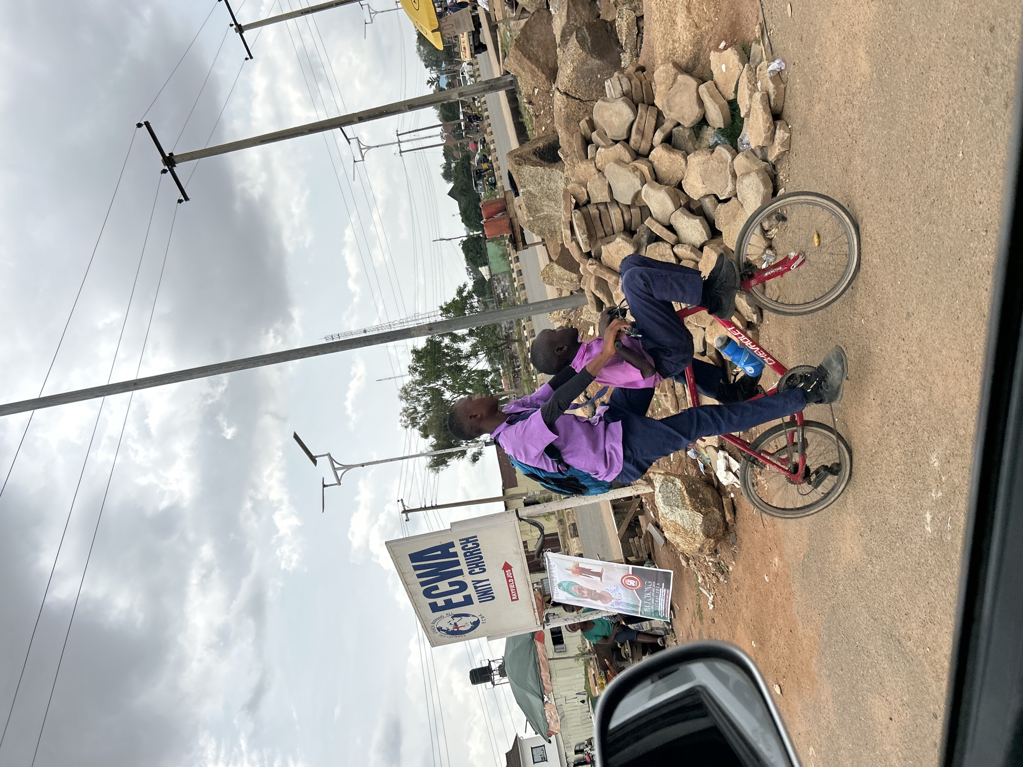 Two children are riding a red bicycle on a paved roadside in Bar Jirgi Summa, Nigeria. One child is on the seat and pedaling, and the other is standing on the bicycle's handlebars, holding onto the rider. The children are wearing purple shirts and dark pants. Behind them is a pile of stacked concrete blocks, and behind that is a road with vehicular traffic and power poles. There is an ECWA Unity Church sign adjacent to the children. The sky is overcast. A portion of a vehicle's side mirror is visible in the lower left corner of the image, indicating the photo was taken from inside a vehicle.