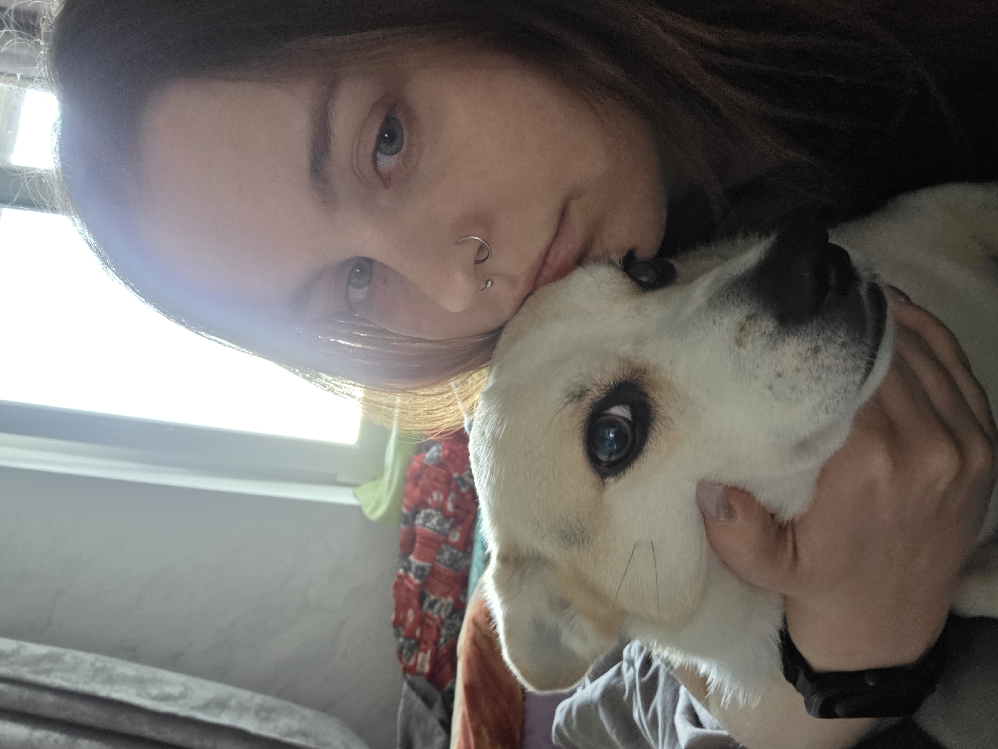 A woman with brown hair and light blue eyes is holding a white and cream-colored dog. The woman has a piercing in her nose. The dog's left eye is visible and appears to be light blue. The woman's right hand is holding the dog's face, and her left hand is unseen. A window is visible in the background, along with a pile of clothes draped over a surface. The photo appears to be taken indoors, possibly in a residential setting.