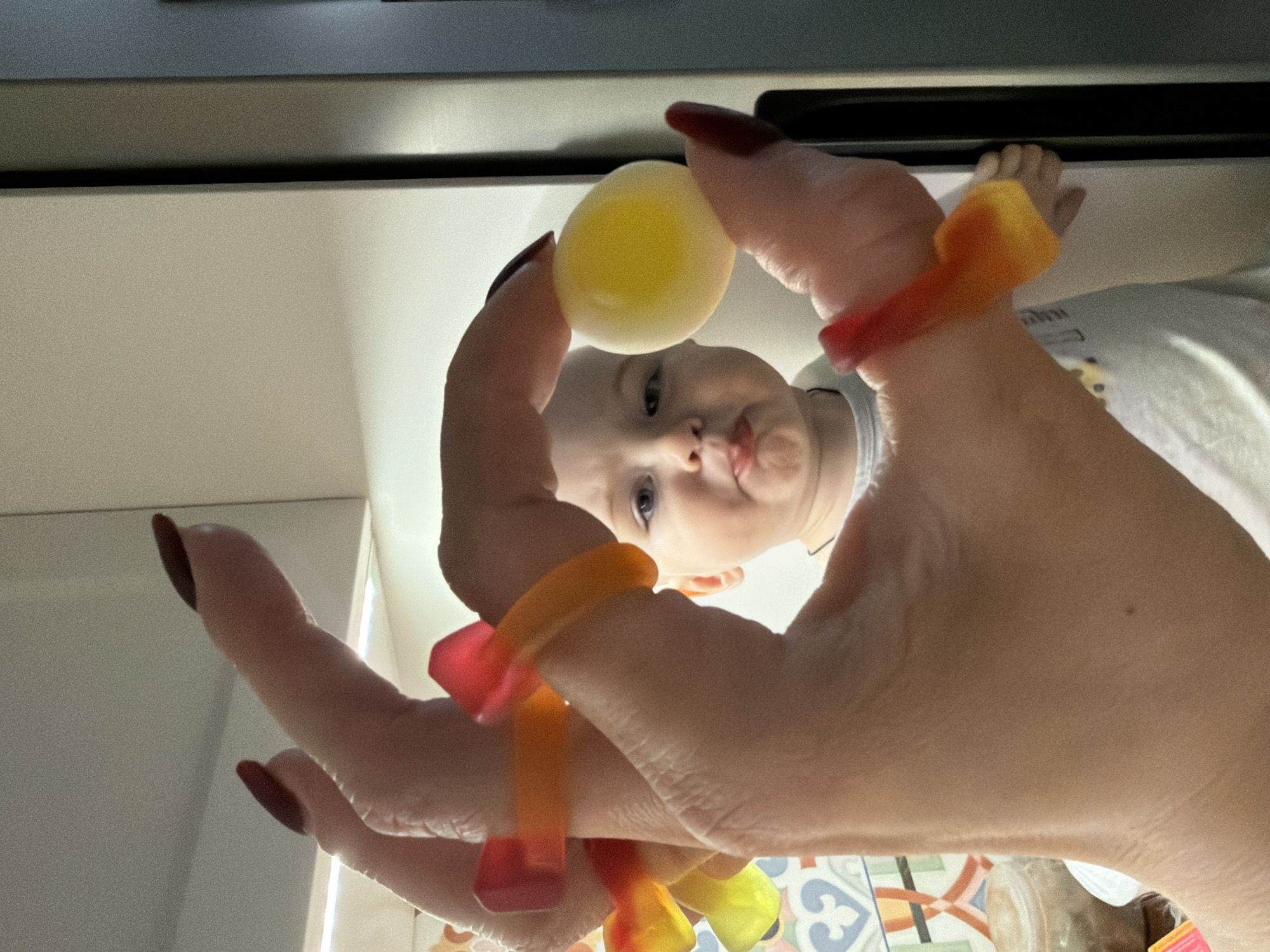 A low-angle close-up captures a baby looking up at a yellow ball held between a person's fingers. The person's hand is adorned with orange and red gummy candies around the wrist and fingers. The person has long, dark red nails. In the background, there is a white cabinet with a gray upper cabinet. The location cannot be confirmed as Votkinsk, Russia.