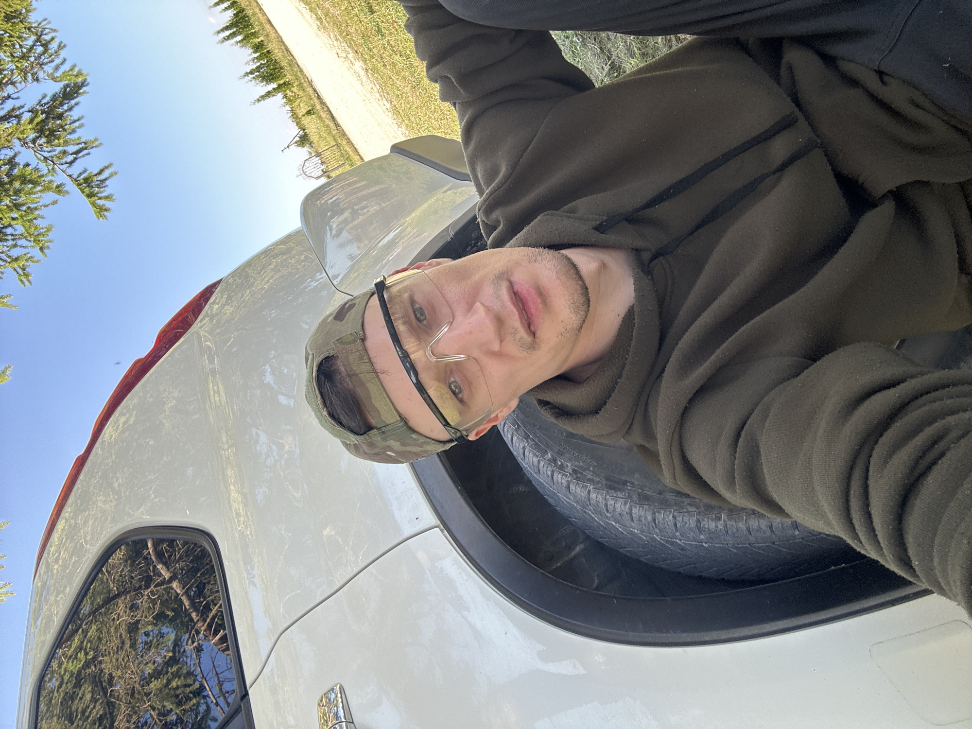 A man is pictured outdoors, leaning against the rear wheel of a white car. He is wearing a camouflage baseball cap and clear safety glasses. His clothing includes a dark green hooded sweatshirt and dark pants. The car appears to be parked on a dirt or gravel surface, with dry grass and some trees visible in the background. The sky is clear and blue. The location is identified as Makiv, Ukraine.