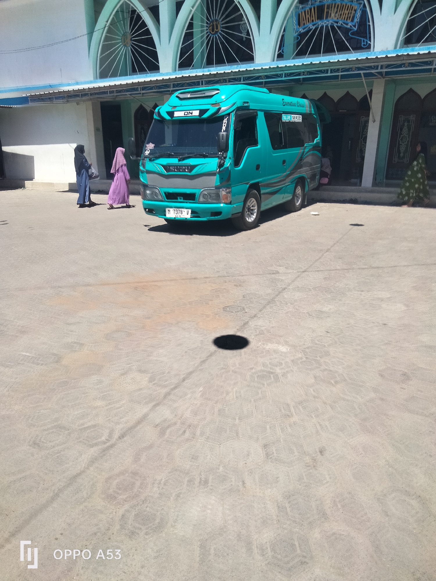 The image captures a bright, sunny day in what appears to be an outdoor area in front of a building with distinctive arched windows. A turquoise colored mini-bus is parked prominently in the foreground, facing towards the viewer. The bus has "ON" written on its front, and "Executive Class" and "BLUE SERIES" on its side, suggesting it is a passenger transport vehicle. A license plate on the front of the bus reads "M 7378 V".
To the left of the bus, two women in traditional Islamic attire (hijabs) are walking away from the viewer. One is wearing a dark blue hijab and dress, and the other is in a light purple hijab and dress. Another woman, dressed in a green patterned garment, is seen walking near the entrance of the building on the right side of the image.
The ground is paved with hexagonal concrete blocks, showing a textured surface. Shadows of the bus and possibly other unseen objects are cast on the ground, indicating the high position of the sun. The building in the background features light blue arched architectural elements and is likely a mosque given the Islamic attire of the people and the style of the building. The text above the entrance appears to be "JABAL RHODOH". The presence of the bus and the building suggest a public or religious gathering place. The overall scene is one of everyday activity in a warm, sunny climate, possibly in a city like Badung, Indonesia.