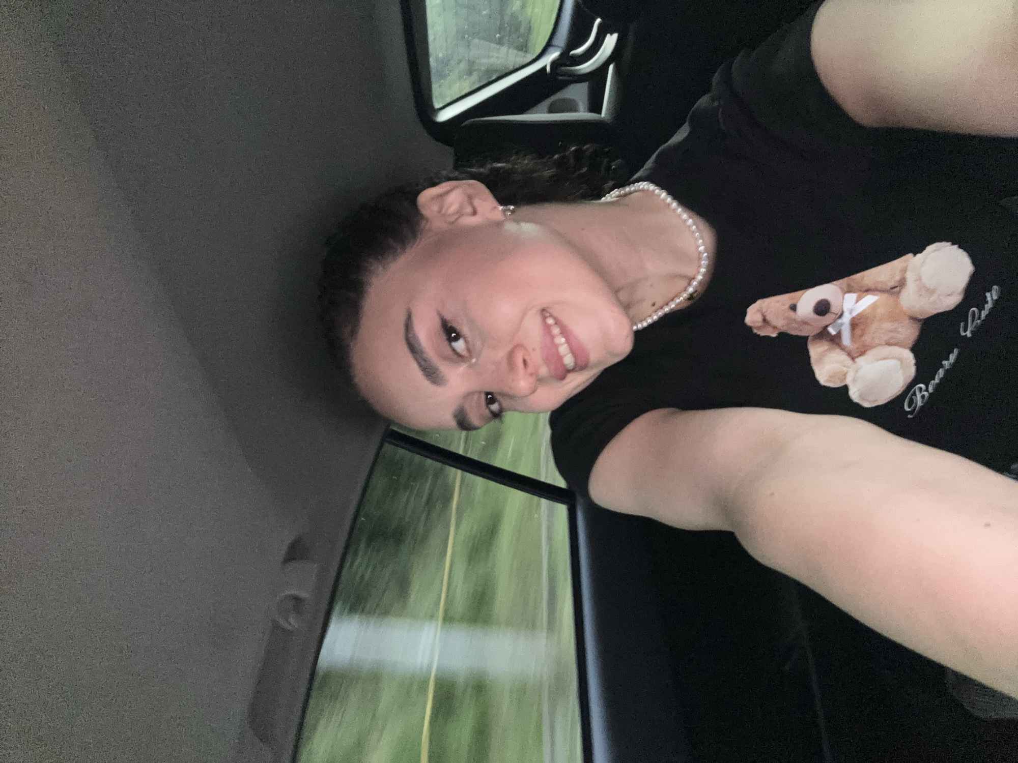 A fair-skinned person with curly dark hair is positioned inside a moving vehicle. The person is smiling. They are wearing a black t-shirt with a teddy bear print, a pearl necklace, and earrings. The background, visible through the car window, suggests an outdoor environment with greenery. The interior of the car includes the ceiling, window, and door panel. There is no evidence to specify that the location is in Tbilisi, Georgia.