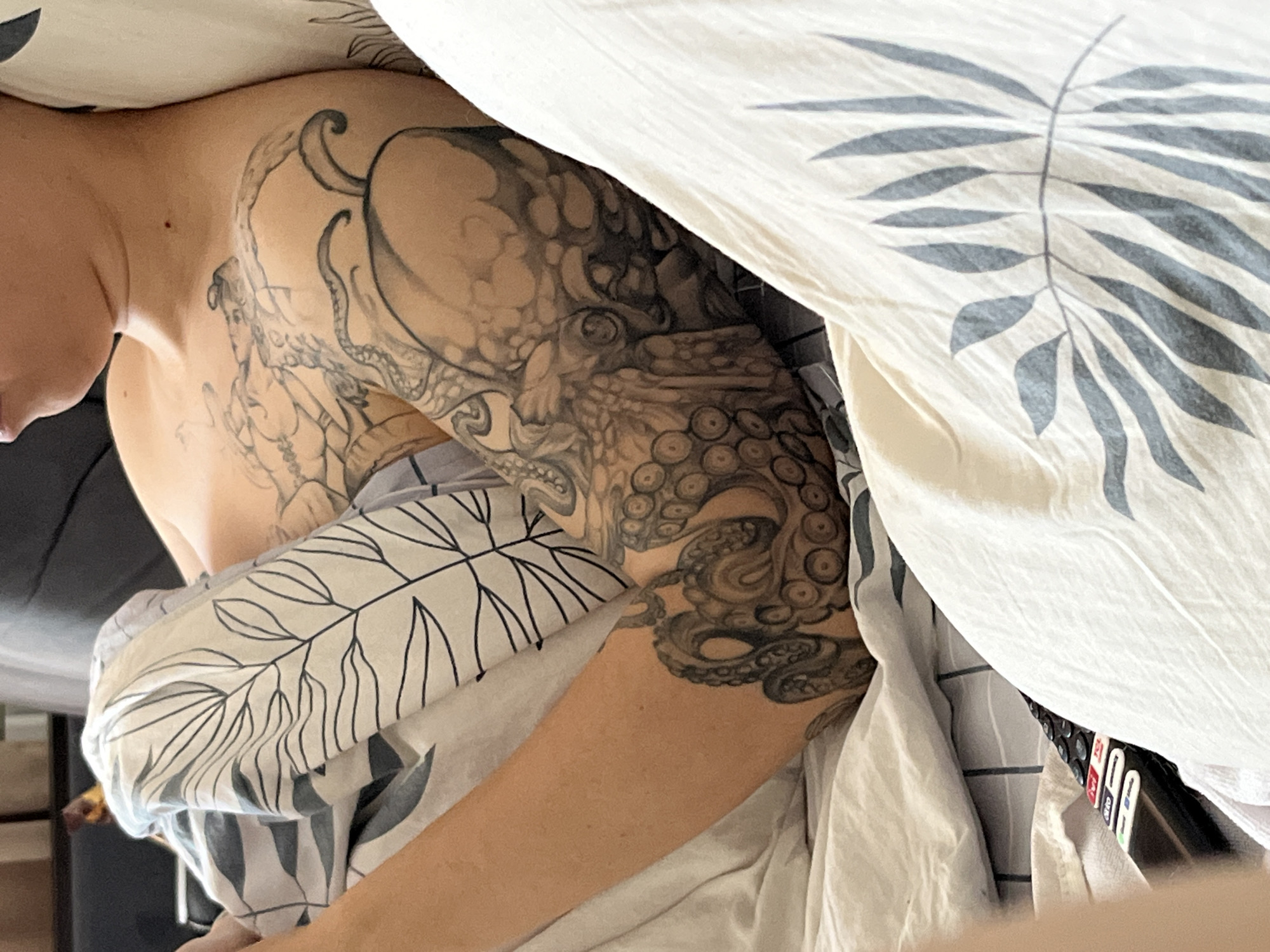 The image shows a person lying in bed. The person has tattoos on their shoulder and arm, including an octopus design. The bed is covered with patterned sheets and a duvet featuring leaf-like patterns. Part of a remote control is visible near the bottom right corner of the frame, partially obscured by the bedding. There's no indication the photo was taken in Chaikovsky, Russia.