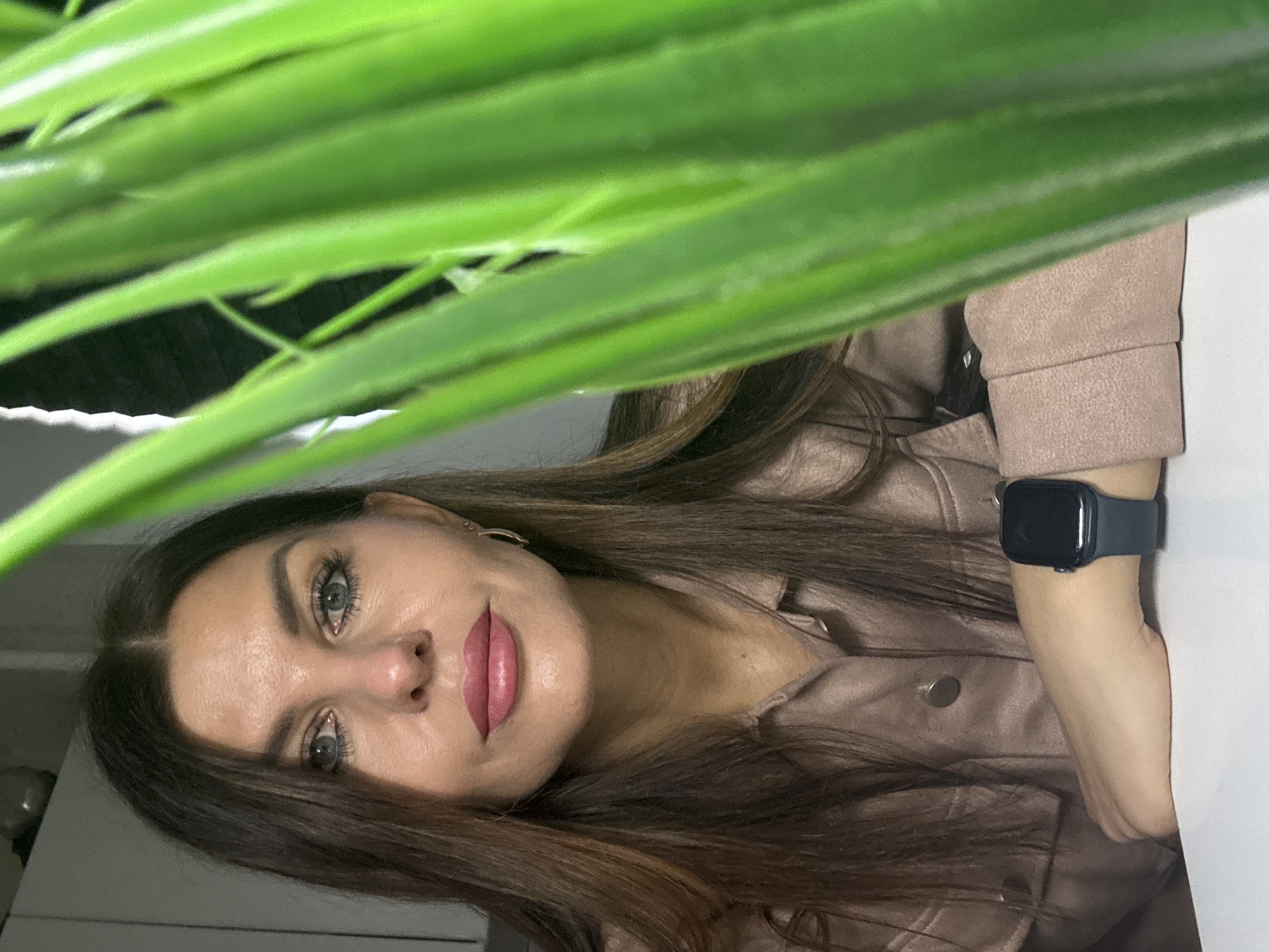 A woman with long, brown hair, wearing a light brown jacket and a dark blue smartwatch, is partially obscured by a plant with long, green leaves. The woman is fair-skinned with blue eyes and pink lipstick. She is looking directly at the camera. The background is blurry, but suggests an interior space. The location is Chaikovsky, Russia.