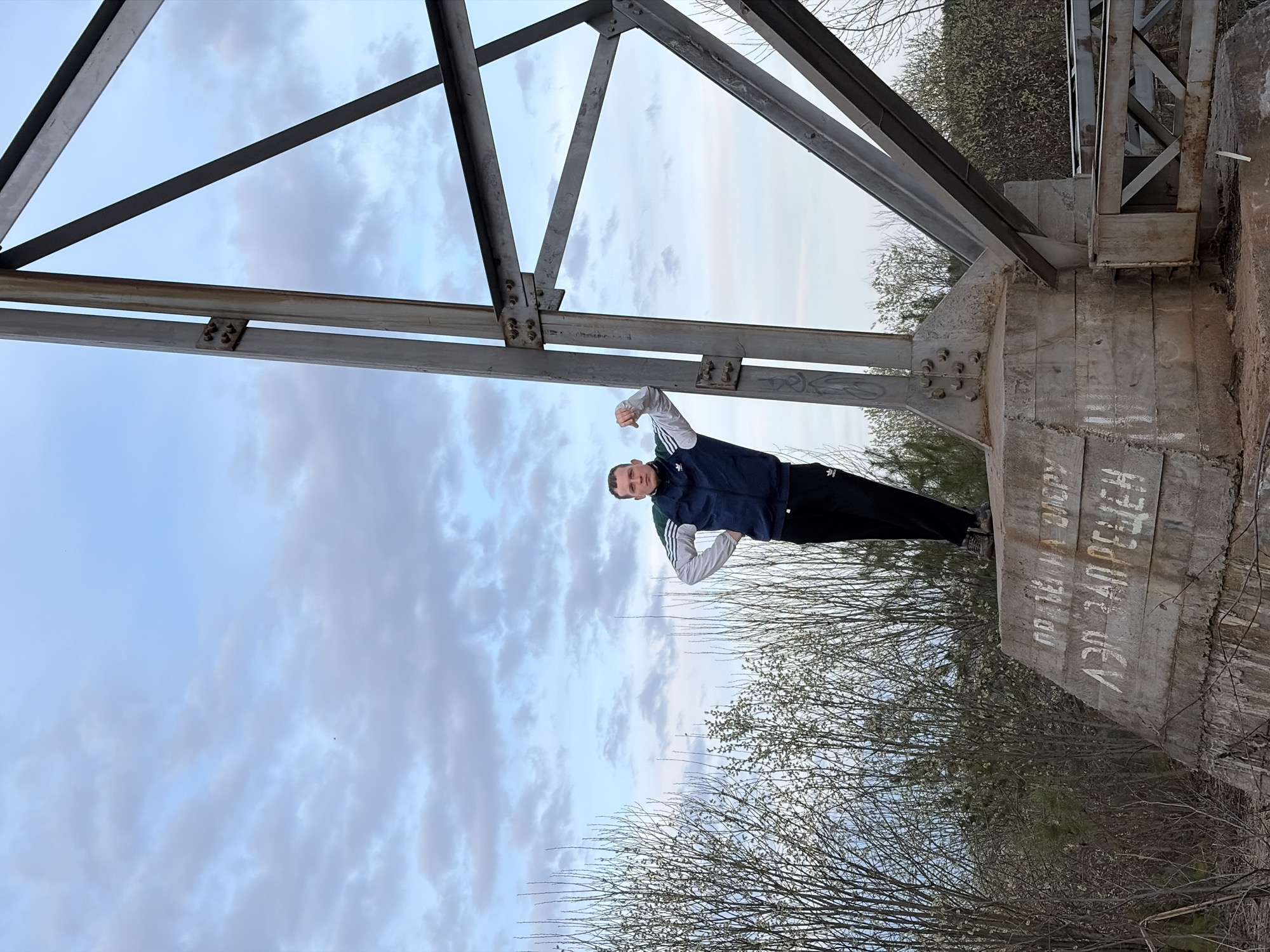 A medium-shot image features a man standing on a concrete structure, seemingly part of a bridge, near a metallic framework. The man is wearing a dark blue jacket with white stripes on the sleeves, and dark pants. He is facing the camera with a slight smile, his hands raised in a gesture of triumph or strength. The concrete structure is weathered and features Cyrillic script painted in white. Bare trees and branches are visible in the background, along with a cloudy sky. The photo appears to be taken from a low angle, looking upwards towards the man and the sky. The location is most likely near or on a bridge support structure in Chaykovsky, Russia, suggested by the Cyrillic script.