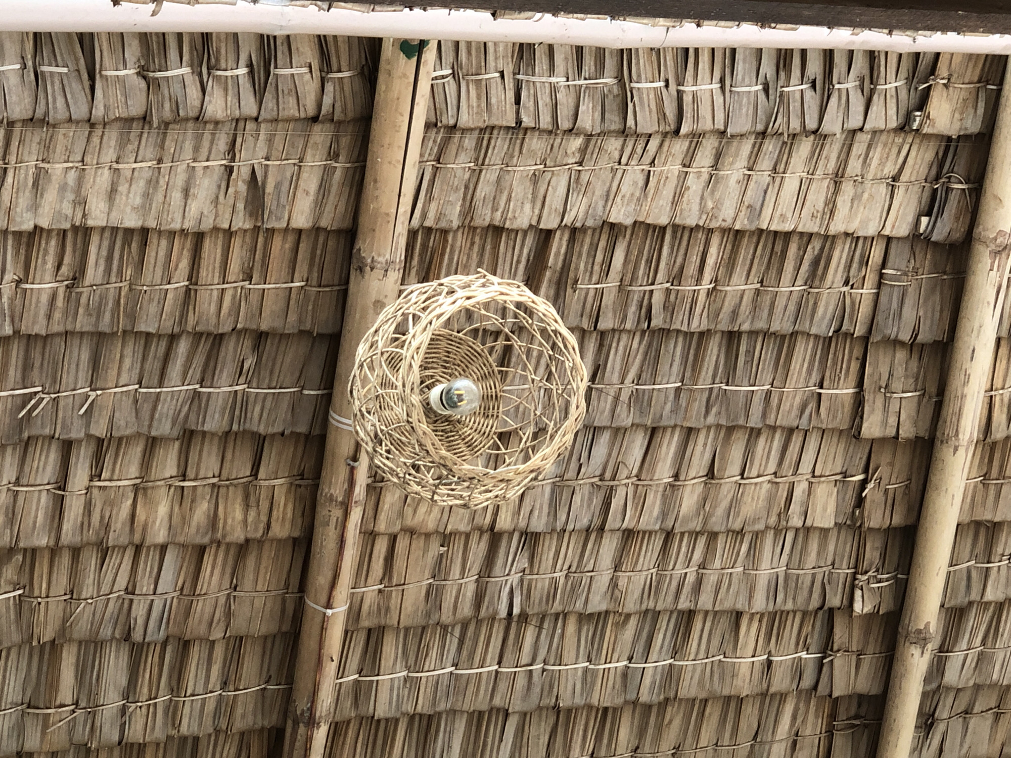 The image shows a close-up of a thatched roof constructed from dried palm fronds, with a woven rattan light fixture hanging from the ceiling. The fronds are layered and secured with white twine, creating a textured pattern. A bamboo beam runs horizontally across the frame, supporting the roof structure. The light fixture is spherical and has a visible incandescent bulb. The overall impression is of a rustic, natural aesthetic, typical of accommodations found in tropical or rural settings. The location is identified as Uteungeulinggang, Indonesia, a place known for its natural beauty and traditional architecture. The scene is static and devoid of any human presence.