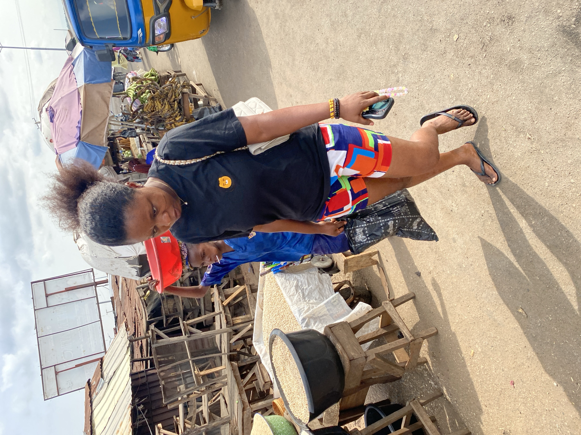 A medium eye-level shot shows a woman in black top and multicolored shorts walking on a street at an open-air market in Lokoja, Nigeria. She is holding a cell phone and a black shopping bag. Adjacent to the street market vendors sit behind sacks and bowls of grains. One vendor, a child, is wearing a blue t-shirt. A makeshift metal and wood structure sits behind the grain stands. An open-air taxi, or Keke, is parked on the street near bananas and other produce.