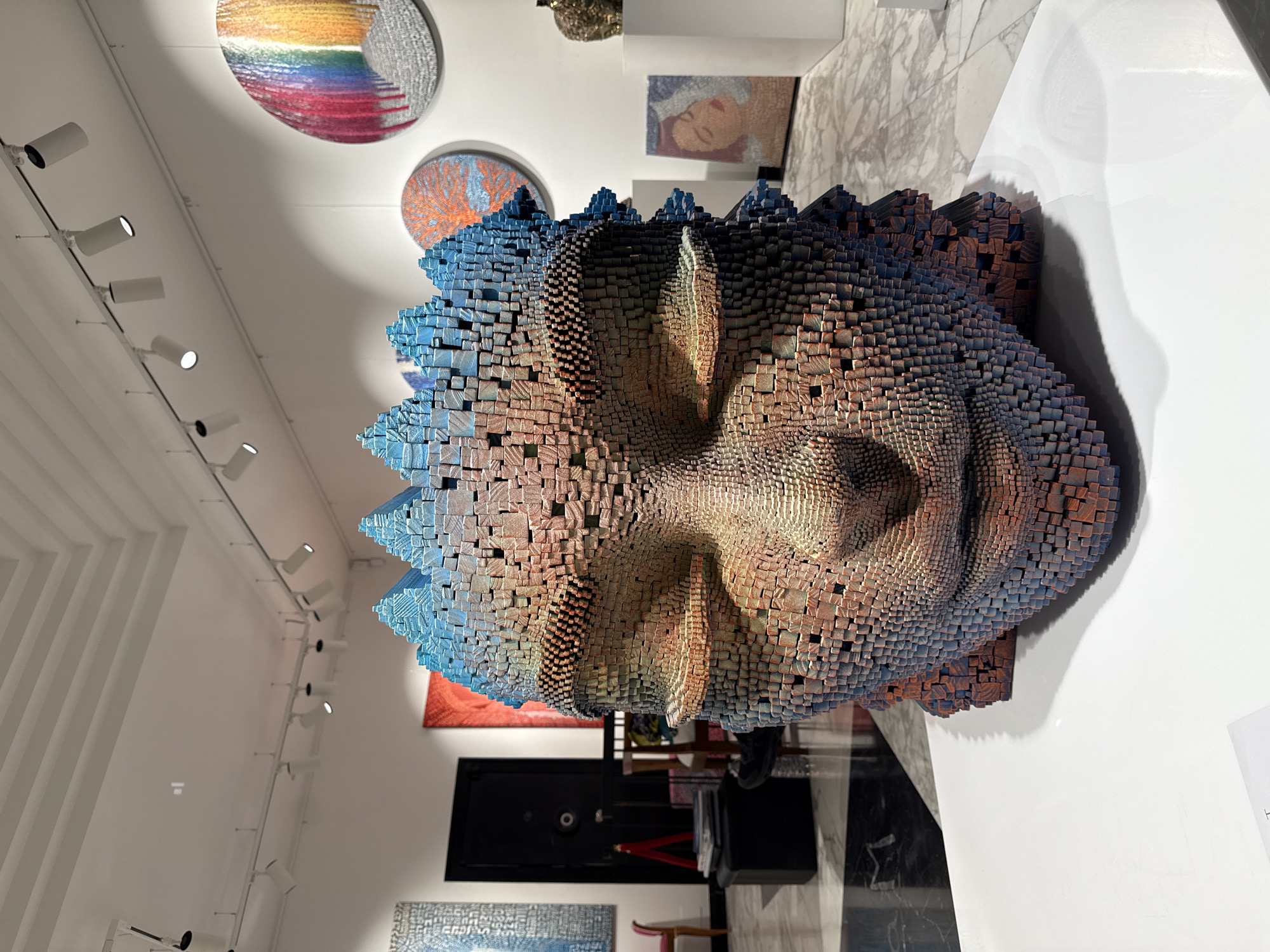 The image captures an interior view of an art gallery in Milan, Italy. The dominant feature is a large, three-dimensional sculpture of a face, possibly a Buddha, constructed from numerous small, colorful cubes. The color palette transitions from blue on the perimeter to warmer tones around the facial features. Above the sculpture, the gallery ceiling is adorned with various circular artworks. Spotlights line the ceiling, illuminating the displays. The gallery's flooring is a polished, black and white marble. In the background, one can discern other art pieces, including paintings. A portion of a black cabinet is also visible. A white placard is next to the sculpture.