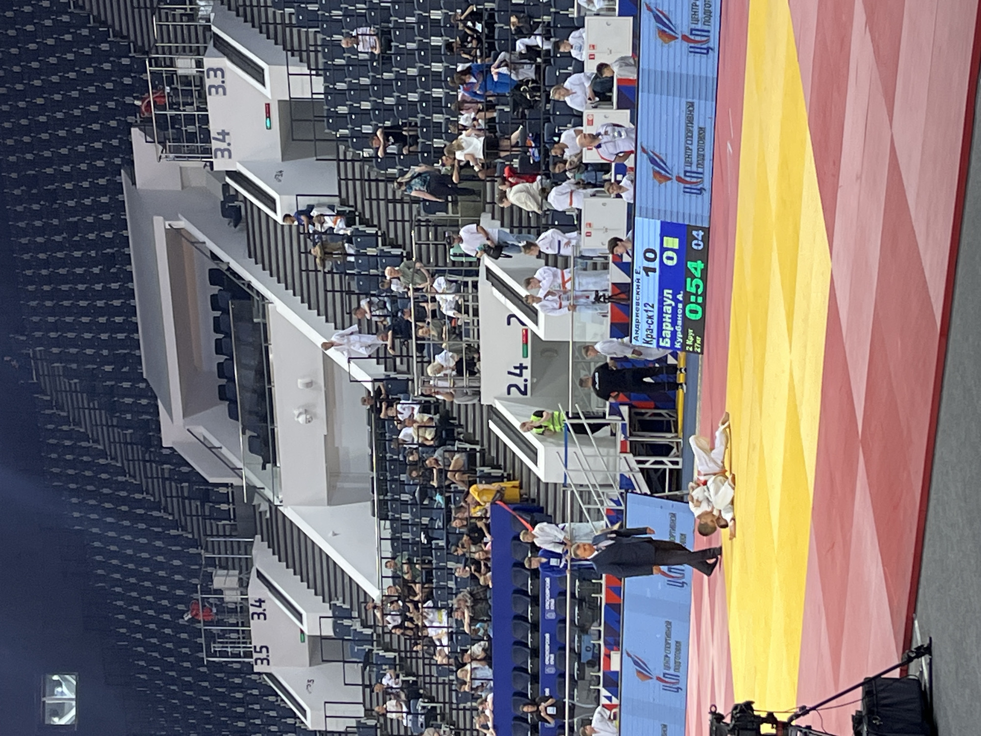 An indoor sports arena hosts a judo competition, viewed from an elevated perspective. The main focus is a vibrant red and yellow judo mat where two young athletes in white judogi are engaged in a match. One judoka appears to be on the ground, while the other stands over them, potentially having just completed a technique or preparing for a hold. A referee in a dark suit stands nearby, observing the action intently.
Adjacent to the mat, a large digital scoreboard prominently displays the match details. It shows "Андриевский Е." (Andrievsky E.) from "Кра-ск 12" with a score of 10, competing against "Курбанов А." (Kurbanov A.) from "Барнаул" (Barnaul) with a score of 0. The time remaining in the match is "0:54", indicating it is the "2 Круг" (2nd Round) for the "27 кг" (27 kg) weight category, with a match number "04".
Behind the mat and scoreboard are tiered spectator stands, mostly dark blue and largely empty, though scattered individuals and groups watch the event. Some spectators are also seen wearing judogi, suggesting they might be fellow competitors or coaches. Staircases providing access to different sections are visible, marked with numbers such as "3.3", "3.4", "3.5", and "2.4". Banners along the arena walls feature the text "ЦЕНТР СПОРТИВНОЙ ПОДГОТОВКИ" (Center for Sports Training), reinforcing the setting as a sports facility in Krasnoyarsk, Russia. Camera equipment is visible in the foreground, indicating professional coverage of the event.