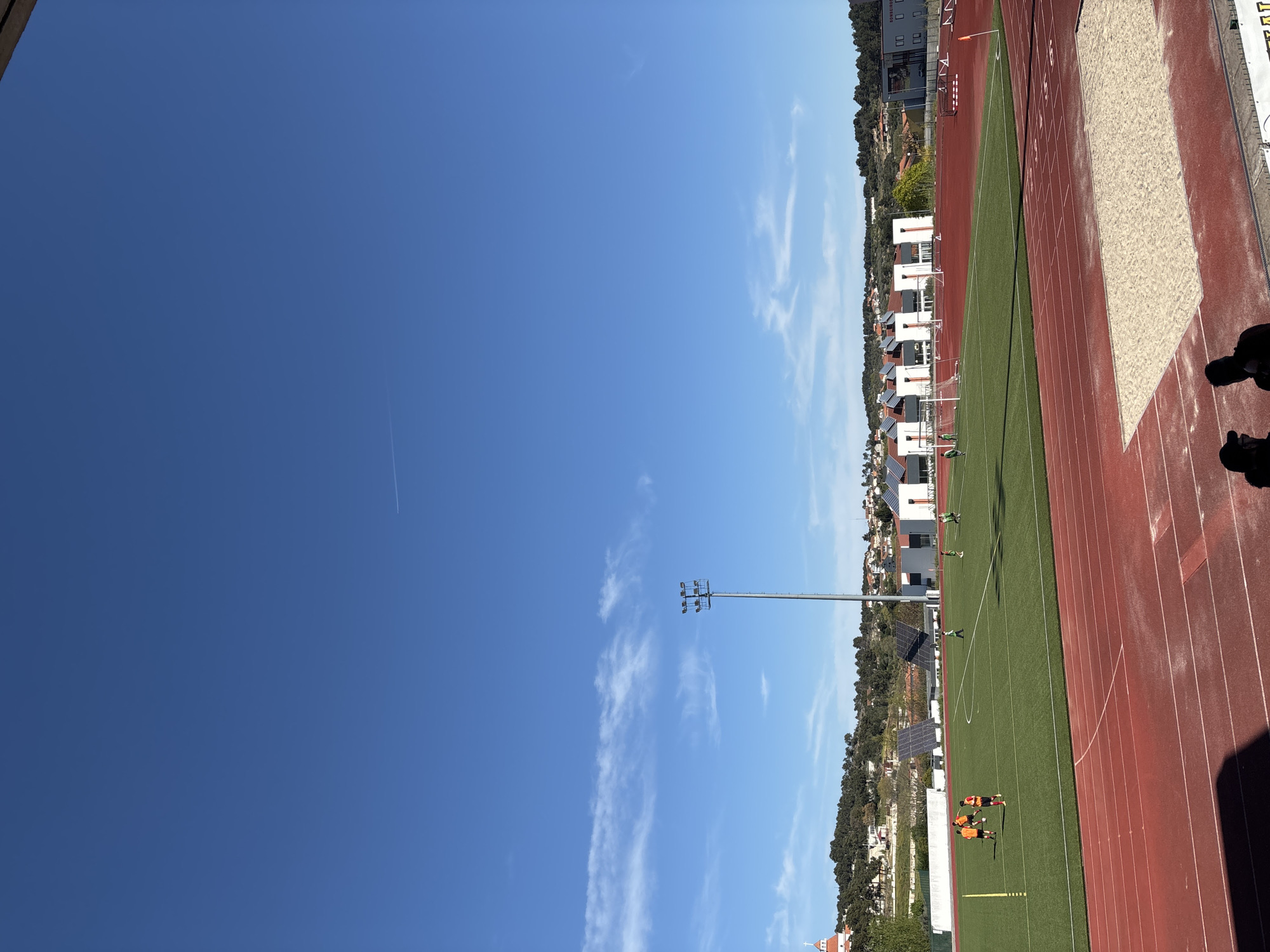 The image shows a partially visible sports complex in Valpaços, Portugal. The sky is mostly clear with a few wispy clouds and a contrail from an aircraft. A tall floodlight pole stands near a running track. The track is red with white lane markers and borders a green field. Several people, dressed in orange and red uniforms, are on the field. White buildings with solar panels are visible in the background, nestled among trees. The edge of a sand pit is also visible.