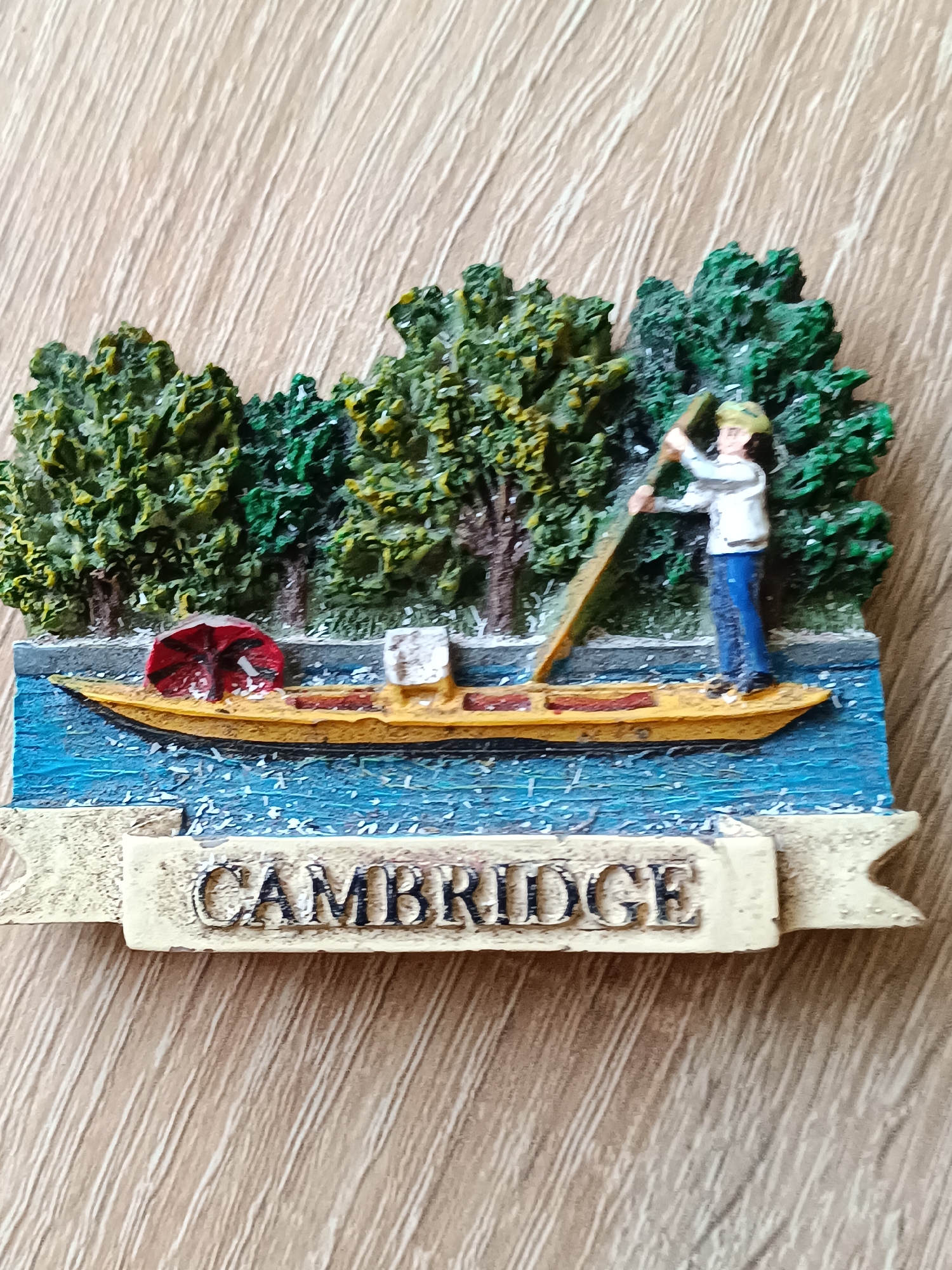 This is a close-up, top-down view of a souvenir magnet. The magnet depicts a miniature scene of a person punting a boat on a river, with trees lining the riverbank and the word "CAMBRIDGE" inscribed on a banner below.
The primary subject is a person, depicted as a male figure wearing a light-colored shirt and blue pants, standing in a yellow boat. He is actively engaged in punting, holding a long pole with both hands and pushing it against the riverbed to propel the boat forward. The boat itself is a narrow, yellow vessel, with a red and black umbrella-like object at its front and a white rectangular object towards the stern.
The scene is set on a blue river, with white flecks representing water splashes or ripples. Behind the river, a dense line of green trees stands, with some trunks visible. The overall impression is a sunny day, indicated by the bright colors and the lack of shadows suggesting overhead sunlight.
The word "CAMBRIDGE" is clearly visible on a light-colored banner running across the bottom of the magnet. The lettering is in a dark, embossed style. The background of the image is a light-colored wooden surface with a prominent grain pattern.
The magnet appears to be a decorative item, likely intended as a souvenir. The scene evokes a peaceful, leisurely activity associated with the location named on the banner. There are no discernible emotions expressed by the figure, as it is a static representation. The time of day and weather are inferred to be fair and bright. The location cue is explicitly provided by the text "CAMBRIDGE". The Tebras, Latvia context does not appear to be visually represented in the magnet itself.