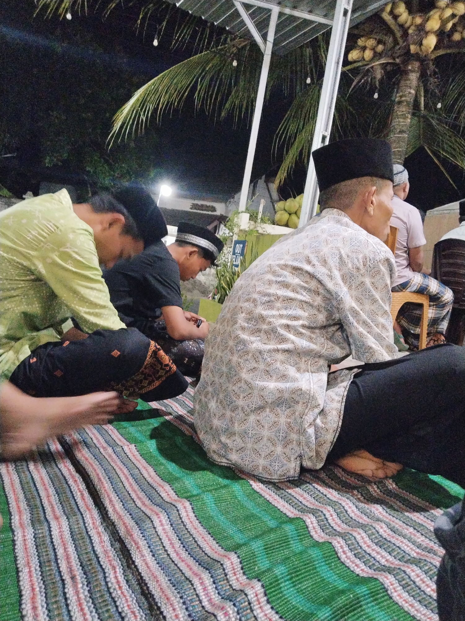 The image depicts an outdoor gathering at night, likely in Prigi, Indonesia, given the presence of a coconut tree. Several individuals are seated on a striped mat on the ground, dressed in traditional attire, including peci (caps) and patterned shirts. One person in the foreground wears a light-colored, patterned shirt and black pants, facing away from the camera. To their left, another individual in a green shirt and black pants is partially visible, leaning forward. In the background, a young person in a black shirt and patterned pants is also seated. A coconut tree with coconuts is visible in the upper right portion of the image, and the scene is illuminated by artificial lights. The overall atmosphere suggests a communal event or religious gathering.