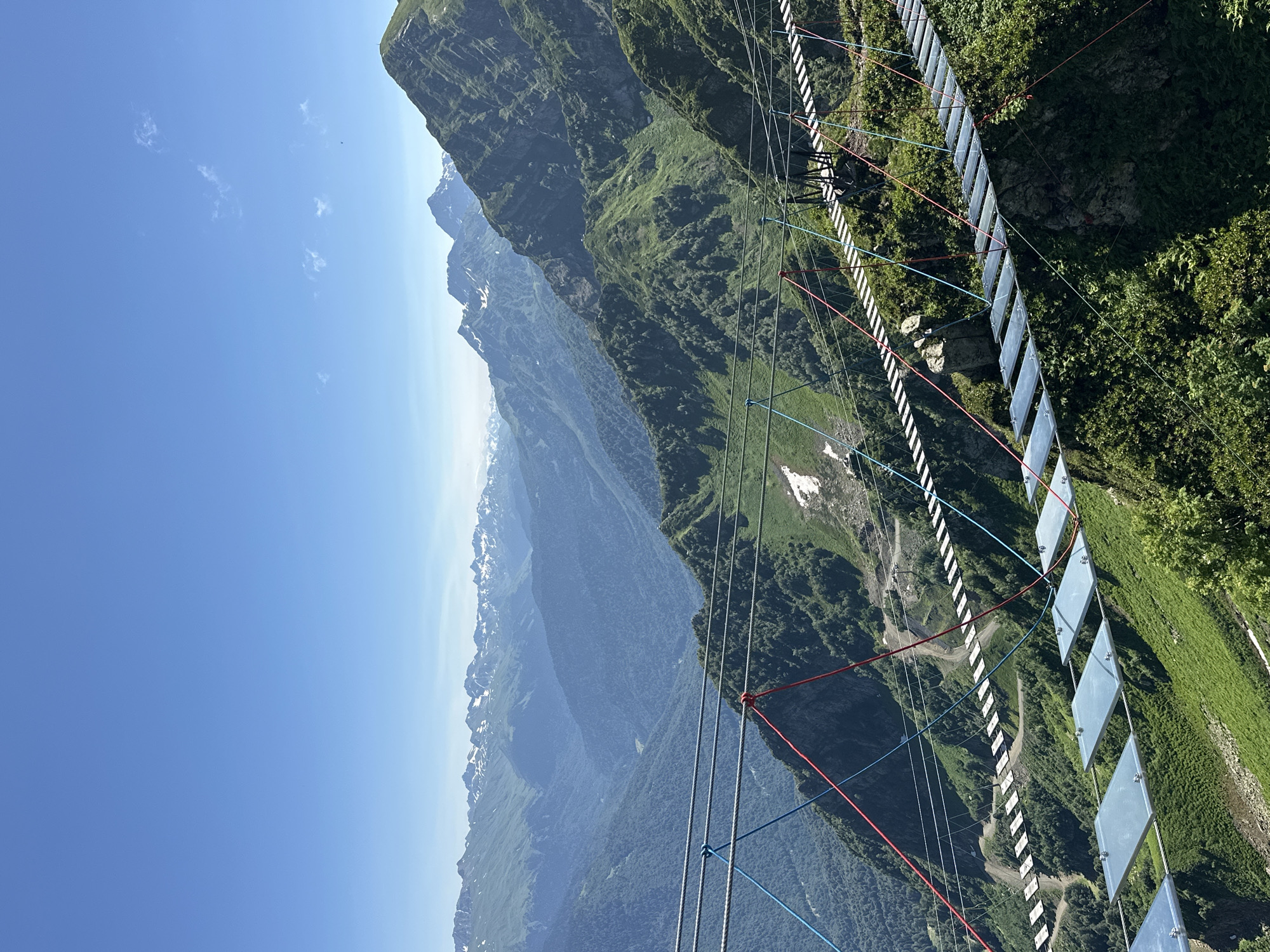 The image depicts a suspended pedestrian bridge, likely part of a mountain resort attraction, in the scenic mountainous region of Estosadok, Russia. The bridge is constructed with metal planks and is supported by thick cables, with colorful red and blue guide ropes adding visual accents. The bridge spans across a steep, verdant mountain slope, with lush green vegetation covering the terrain. In the background, a vast expanse of majestic mountains, some with snow-capped peaks, stretches towards the horizon under a clear, bright blue sky. The overall scene captures a breathtaking natural landscape enhanced by human engineering.