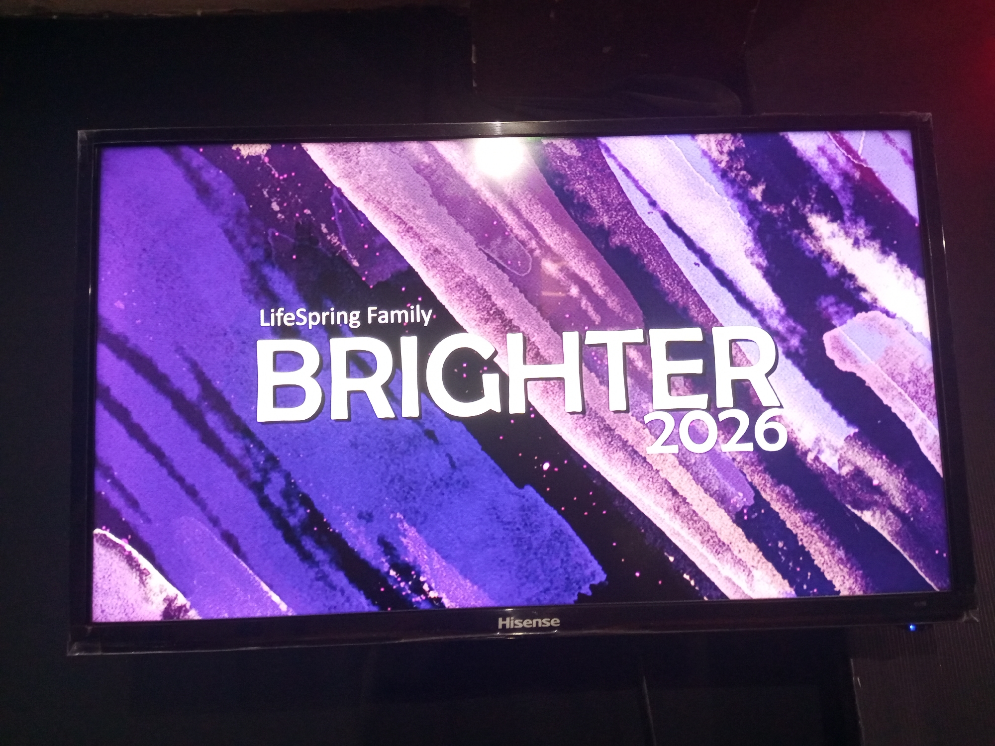 A Hisense television screen displays "LifeSpring Family" above "BRIGHTER" and "2026" in white lettering against a purple, abstract background. The screen is situated in a dimly lit environment. There is no information available that connects the content to Onitsha, Nigeria.