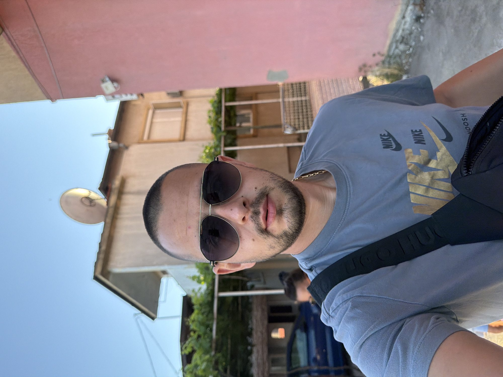 The media file shows a close-up, rotated, portrait-style photograph of a man wearing sunglasses and a light blue t-shirt with a gold Nike logo and text. He has short dark hair and a goatee. The man is looking towards the camera, with his head tilted slightly.
In the background, a building with a pink facade is visible, along with another building with windows. Green foliage and what appears to be a street or alleyway can also be seen. The sky is a clear, bright blue, suggesting it is daytime. There are hints of urban architecture, consistent with the provided location context of Burgas, Bulgaria. The overall mood of the photograph appears casual and candid. The image orientation suggests it may have been taken with a mobile phone.
