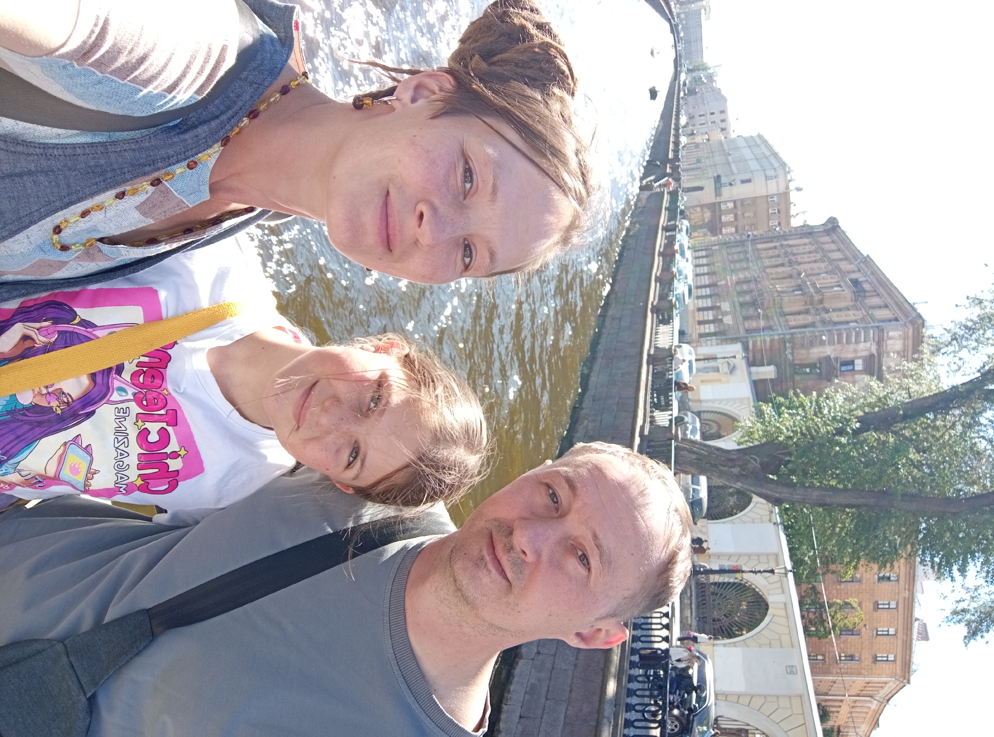 A selfie taken outdoors shows a family of three standing in front of a canal in Saint Petersburg, Russia. The woman in the foreground on the left has her hair in dreadlocks piled on top of her head and is wearing a denim vest over a patterned top and a beaded necklace. The girl in the middle has freckles and is wearing a white t-shirt with a cartoon graphic. The man on the right is wearing a gray sweatshirt and has a strap from a bag across his chest. They are all smiling and looking at the camera. The canal is wide and appears to be lined with a stone embankment. In the background, there are several historic buildings and trees. The water in the canal is reflecting the sunlight, creating a shimmering effect. The architecture of the buildings suggests a location in the historic center of Saint Petersburg, likely along one of the city's many canals, such as the Griboyedov Canal or the Fontanka River.