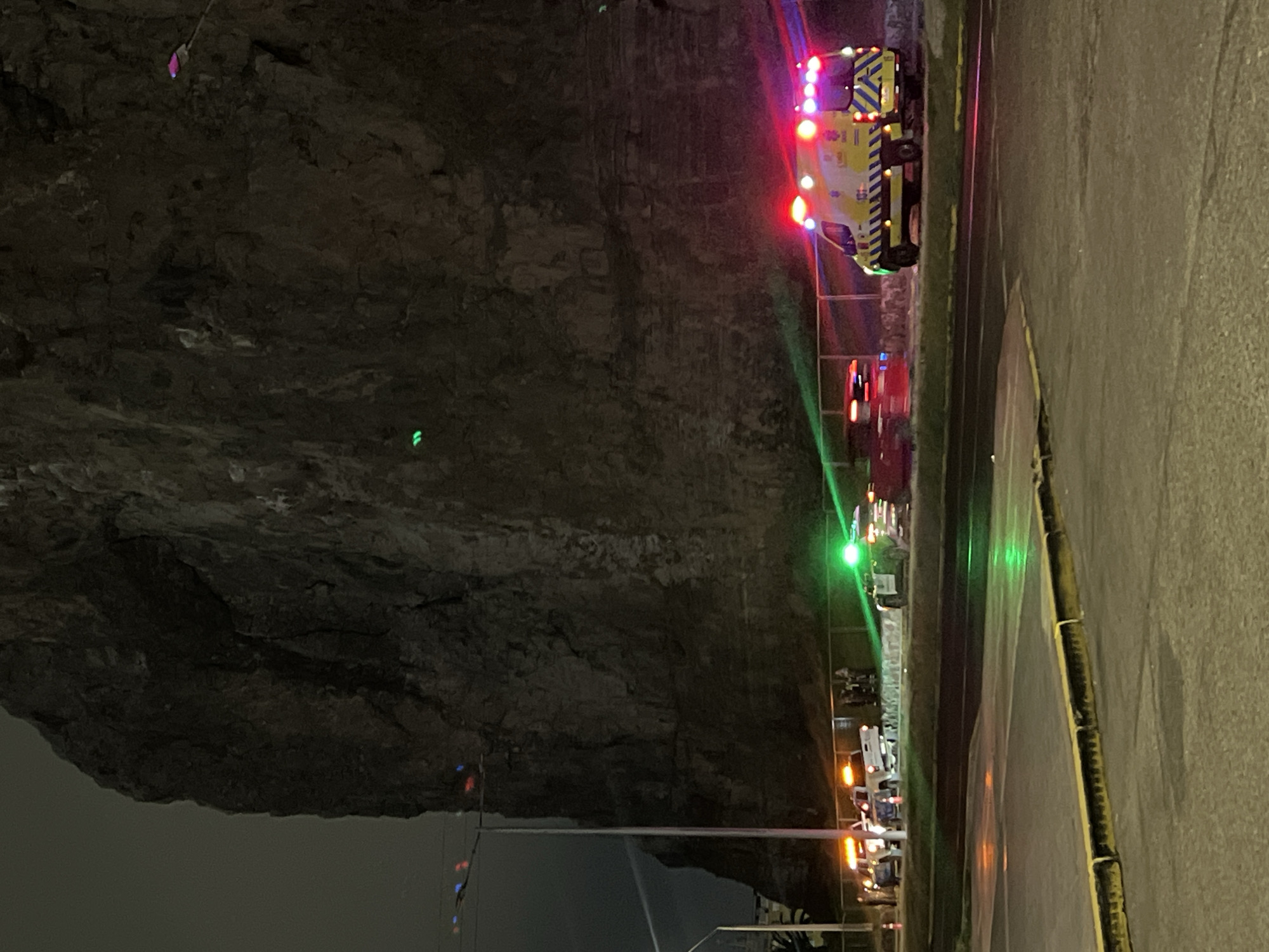 The media file presents a nighttime outdoor scene, likely in Arica, Chile, depicting an emergency situation. A large, dark rocky cliff face dominates the left and central parts of the image, appearing rugged and imposing. To the right, an asphalt road with a yellow curb or lane marking is visible.
A prominent white and yellow ambulance, with flashing red, blue, and yellow emergency lights, is positioned on the road. The text "AMBULANCIA" and the number "131" are discernible on its side. Further down the road, other emergency vehicles with flashing orange and red lights are present, suggesting the involvement of additional services like fire or police.
The scene is illuminated by the emergency vehicle lights, along with distinct green laser-like beams or reflections that span across the cliff face and road. These green lights, coupled with a small, isolated pink/purple light visible higher on the cliff, hint at a search or technical rescue operation possibly focused on the cliff face. Faint blue and red lights are also seen in the lower left section, possibly from distant sources or decorative lights. The time of day is clearly night, and the weather appears clear with no precipitation. No people are explicitly visible in the frame, though their presence is implied by the emergency response.