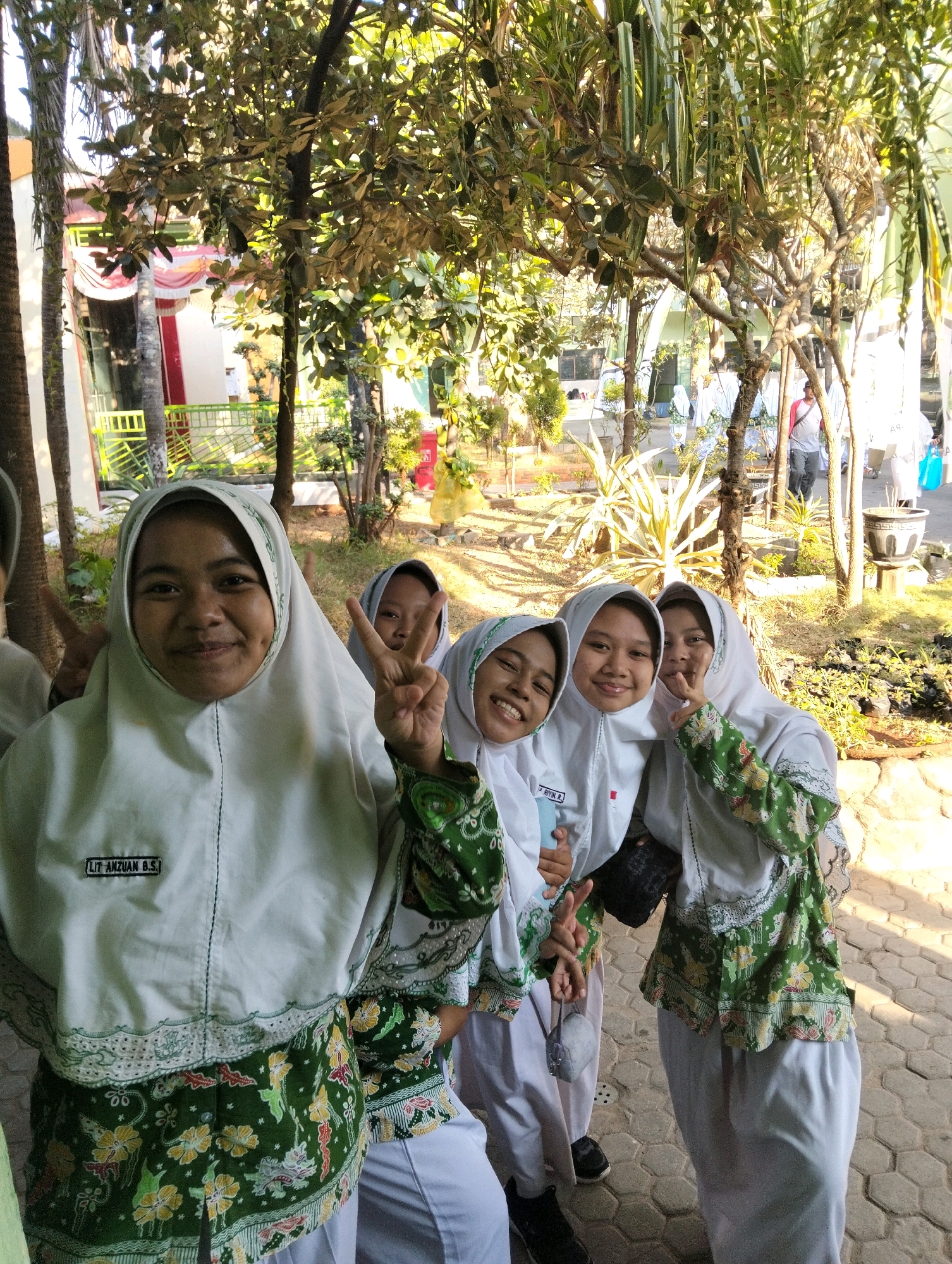 The image depicts four young women, likely students, posing for a photograph outdoors. They are all wearing white headscarves (hijabs) and uniforms that include white tops with green accents and batik-patterned skirts. The woman in the foreground on the left is smiling and making a peace sign with her fingers. The other three women are also smiling and in the background, with two of them also making peace signs. They are standing on a paved walkway with some greenery and trees in the background, suggesting an outdoor school or public space. The lighting indicates it is daytime. The presence of batik patterns on their clothing, and the overall scene, are consistent with locations in Indonesia, and specifically may represent a school in the town of Dagan, Indonesia.