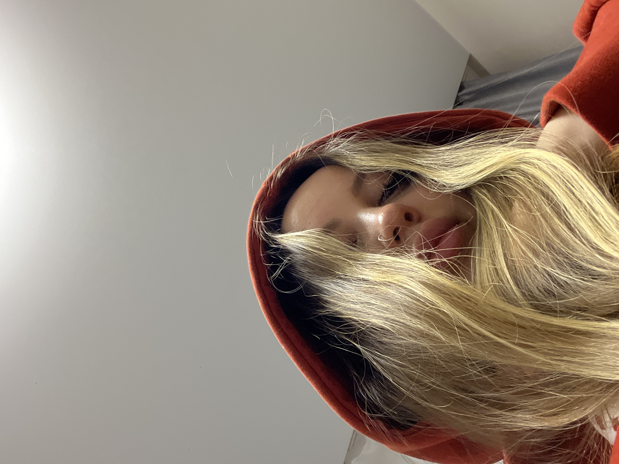 A low-angle close-up photograph depicts a woman with blond hair and fair skin wearing a red hooded sweatshirt. Part of her face is obscured by her hair. The background is a light gray wall. There is no indication of this being Krasnodar, Russia.