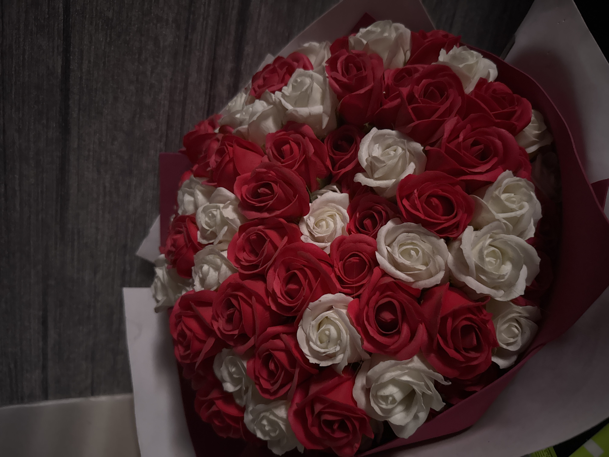 A bouquet of red and white roses is displayed against a dark wooden background. The roses are arranged closely together, forming a full, rounded shape. The bouquet is partially wrapped in pink paper. The lighting is dim, creating shadows on the wooden surface and highlighting the textures of the rose petals. There are no people or other objects visible in the scene. The location is unknown.