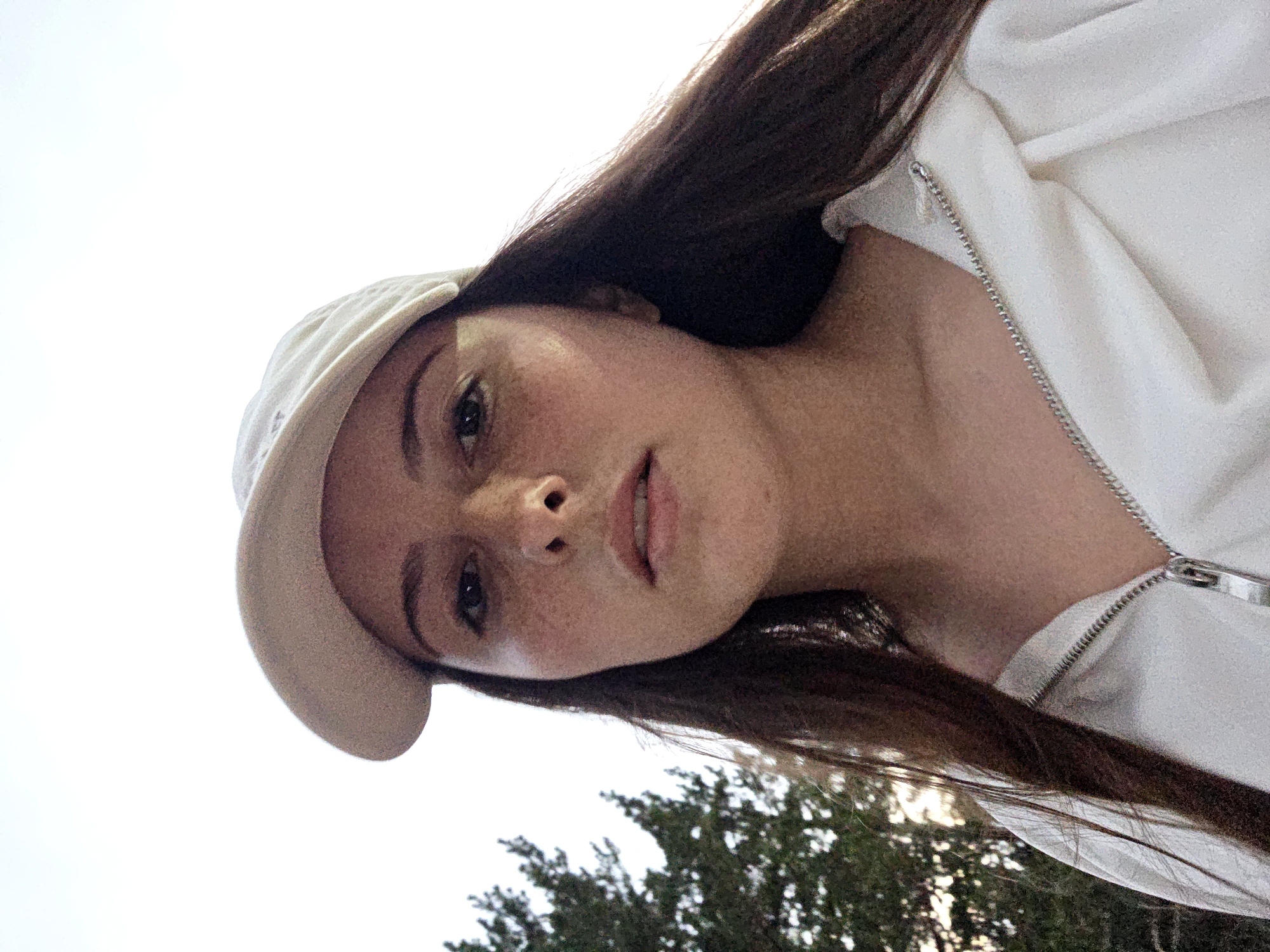A young woman with long, dark brown hair and a light-colored baseball cap is pictured from a low angle, looking directly at the camera. She has fair skin with freckles, dark eyes, and is wearing a white zip-up hoodie. Her face is framed by her hair, which extends over her shoulders. In the background to the left, a dark green evergreen tree is visible against a bright, overcast sky. The lower part of the image shows the zipper and collar of her hoodie.
