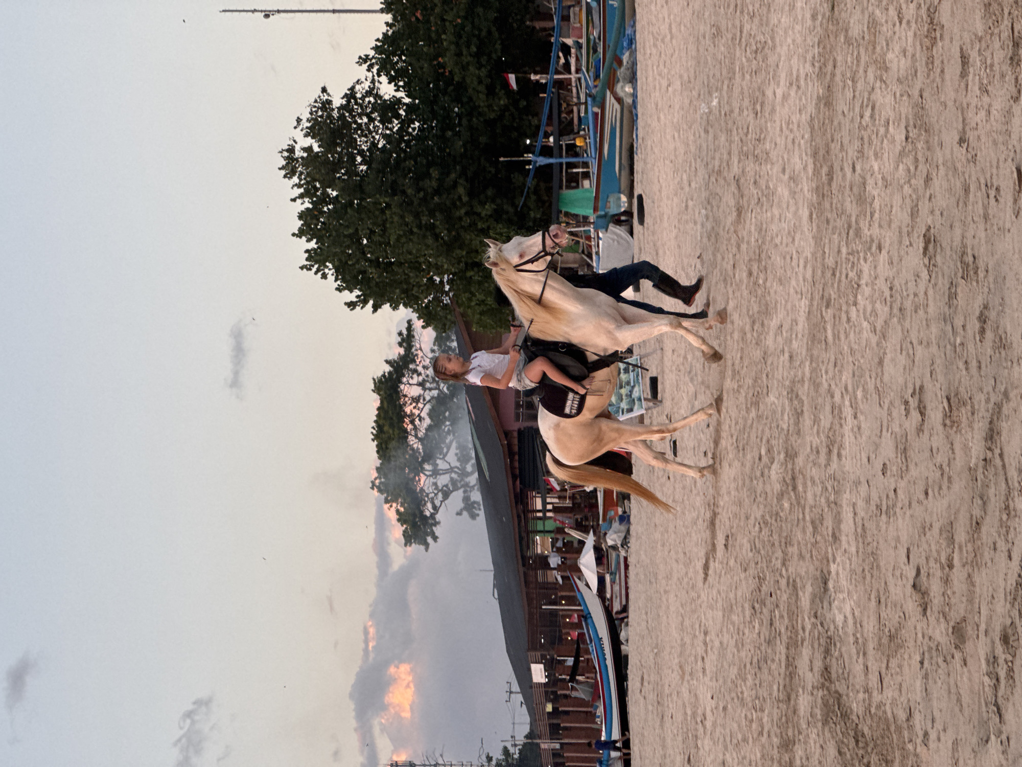 In Kuta Selatan, Indonesia, a young girl with braided blonde hair rides a pale horse along a sandy beach. She wears a white t-shirt and light-colored shorts. The horse is equipped with a black saddle and bridle. A person in dark pants and black boots walks beside the horse, appearing to guide it. In the background, several traditional Indonesian fishing boats are docked on the shore. Behind the boats, a long, open-air structure, possibly a restaurant or market, stands with a dark roof. Lush green trees and a tall communication tower are visible further in the distance. The sky is a soft gradient of pale blue and pink, suggesting either dawn or dusk. The overall atmosphere is serene, with the gentle sounds of the ocean implied by the beach setting.