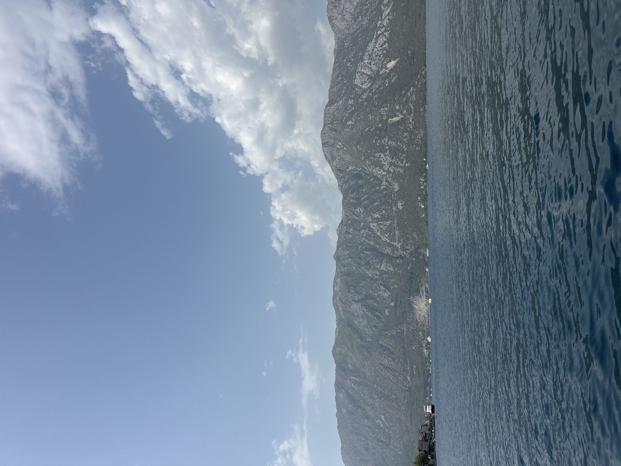 A serene view of the Bay of Kotor, with the mountains of Perast rising majestically in the background. The deep blue water in the foreground is gently rippled, reflecting the clear blue sky dotted with fluffy white clouds. Several boats are visible on the water, some anchored near the shore, while others appear to be sailing. The coastline is dotted with traditional stone buildings, characteristic of the historic town of Perast. The mountains themselves are rugged and covered in sparse vegetation, their slopes illuminated by the bright daylight.