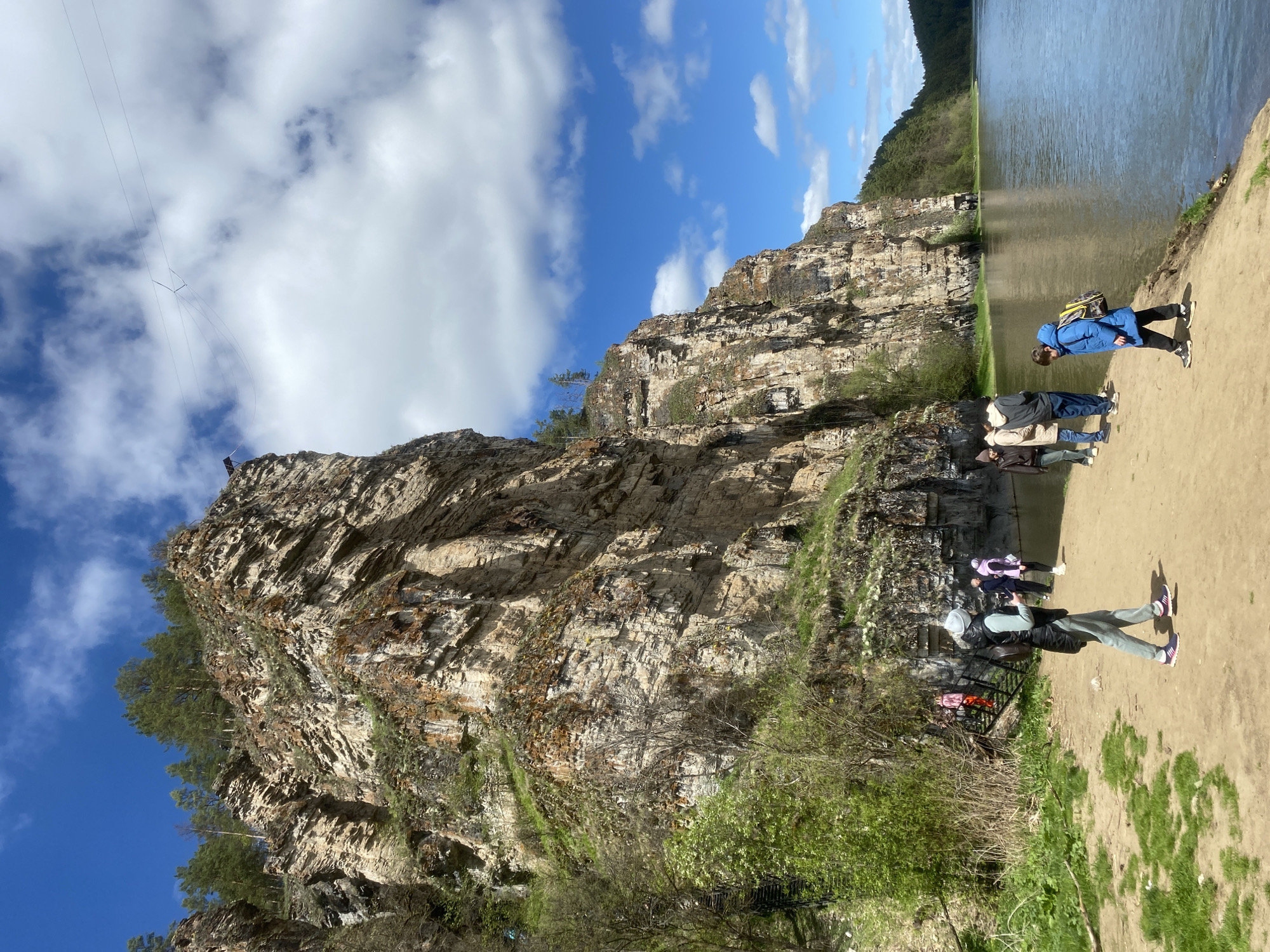 The image depicts a group of people standing along a sandy shore next to a body of water, adjacent to a large, rocky cliff. The cliff is composed of layers of sediment and is partially covered with vegetation. Above the cliff, the sky is partly cloudy with patches of blue. There appears to be a structure or platform on the top of the cliff with a wire or cable extending upward into the sky. The location is Unknown, Russia. The group of people is diverse in attire and appears to be casually interacting with each other.
