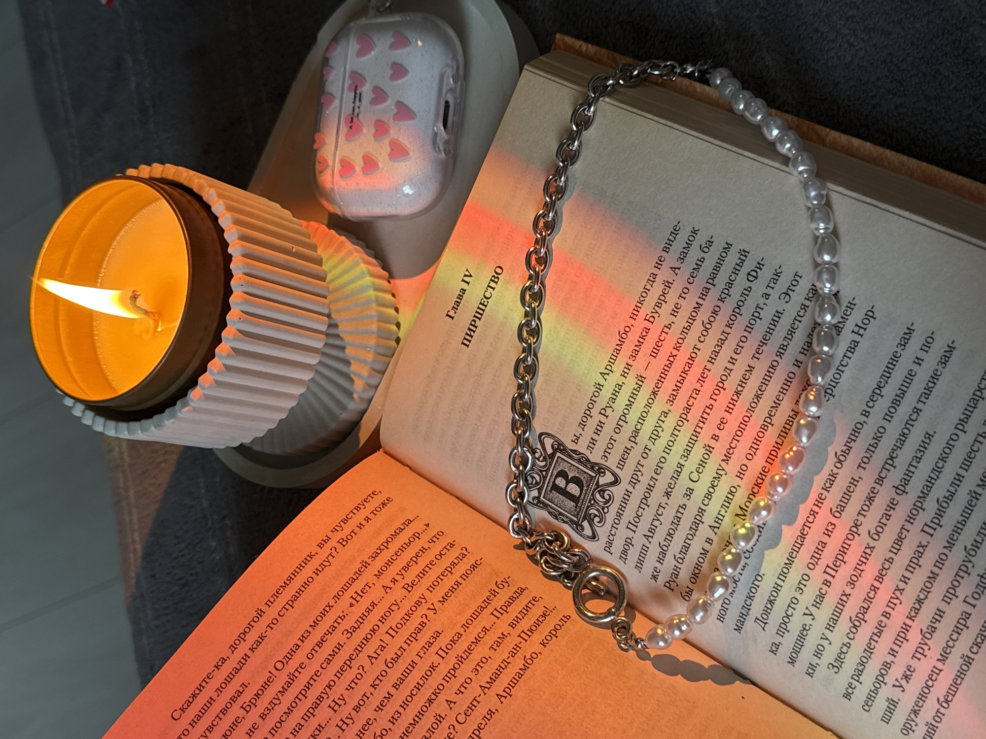 A still life composition is captured in an interior setting in Unknown, Unknown. An open book with Cyrillic text, visible on cream-colored pages, lies horizontally on a dark, textured surface, possibly fabric. The left page displays a dense block of text, while the right page shows "Глава IV" and "ПИРШЕСТВО" at the top, along with a large, ornate letter 'B' within a decorative border. A necklace, featuring a silver-toned chain section and a string of white pearls, is draped across the opened book. The silver chain segment rests near the spine, while the pearl section extends down the right page. A circular clasp with an intertwined design is visible on the chain.
To the upper right of the book, a lit candle in a metal tin is positioned within a white, fluted ceramic holder. This holder and the candle rest on a light-colored, circular tray. To the right of the candle, also on the tray, sits a clear protective case for white AirPods, adorned with multiple small pink heart decals. A faint inscription "A lot can happen in a year" is visible on the AirPods case. The scene is illuminated by the candle's warm glow, which, combined with another light source or optical effect, casts prominent red, orange, yellow, and green light streaks across the book pages and parts of the surrounding objects. The background behind these items is dark and indistinctly textured.
