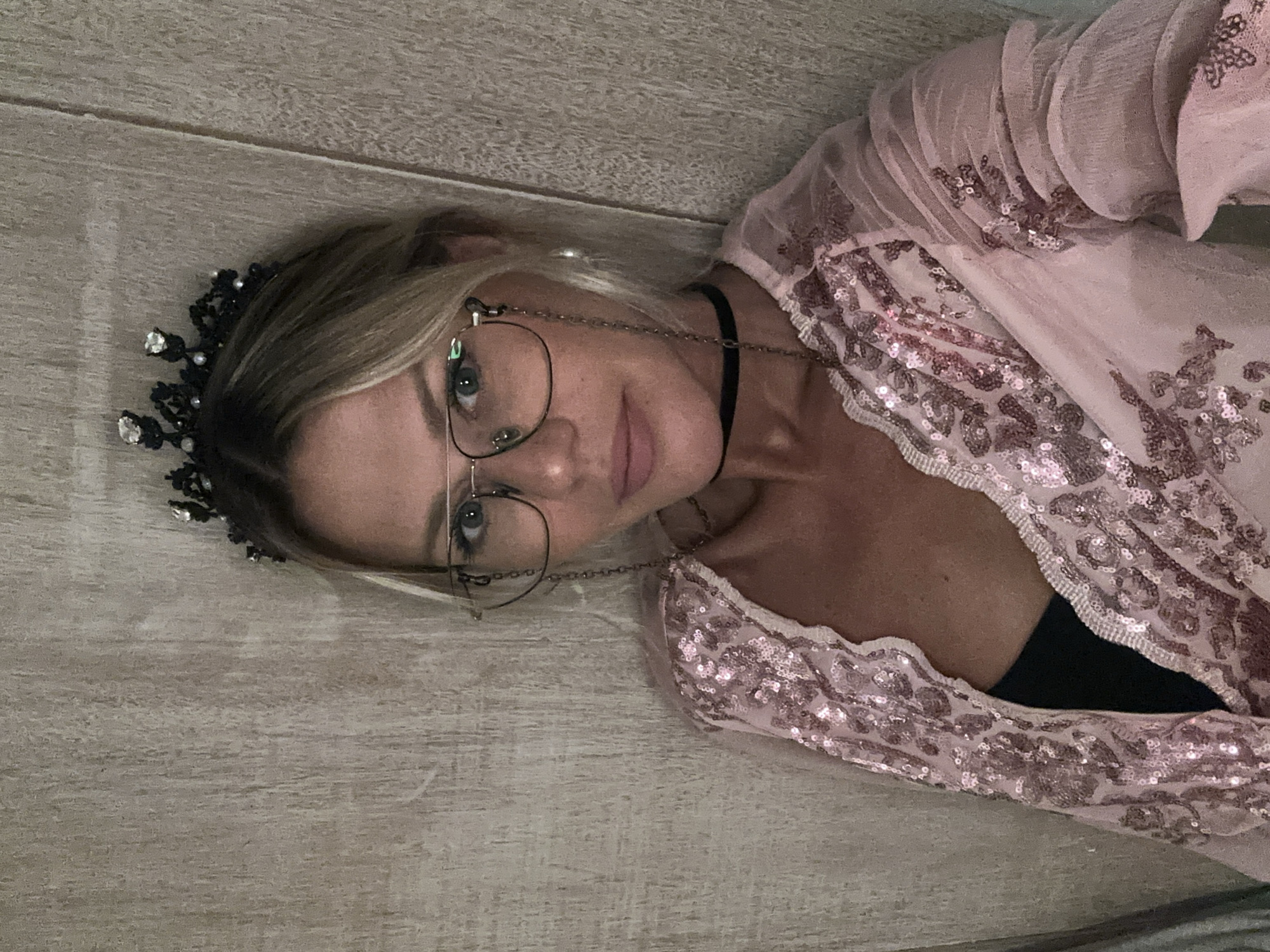 A fair-skinned woman lies on a light brown wooden surface, possibly a floor. She is wearing a pink, sequined top or dress over a black top. She has blonde hair and is wearing glasses with a chain. A black crown-like accessory is on her head. A black choker is around her neck. The location is potentially North Kuta, Indonesia. There are no visible interactions or actions besides the woman lying down. The environment appears to be indoors.