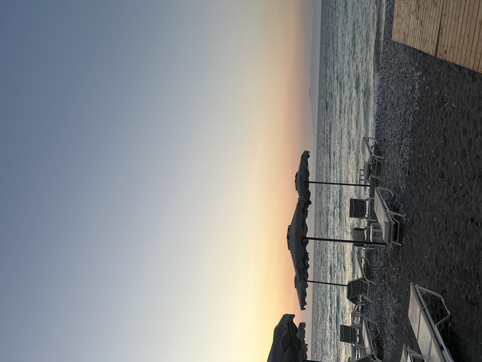 This image captures a serene beach scene in Rhodes, Greece, likely during either sunrise or sunset. The foreground features a dark, pebble-strewn beach with several empty lounge chairs and open umbrellas. A wooden boardwalk runs along the right side of the frame, leading towards the water. The middle ground is dominated by the calm, shimmering sea, with gentle waves lapping at the shore. In the distance, a faint outline of islands or a coastline is visible against the horizon. The sky displays a soft gradient of colors, transitioning from a pale blue at the top to warm hues of orange and yellow near the horizon, indicative of twilight. There are no people or animals present in the scene.