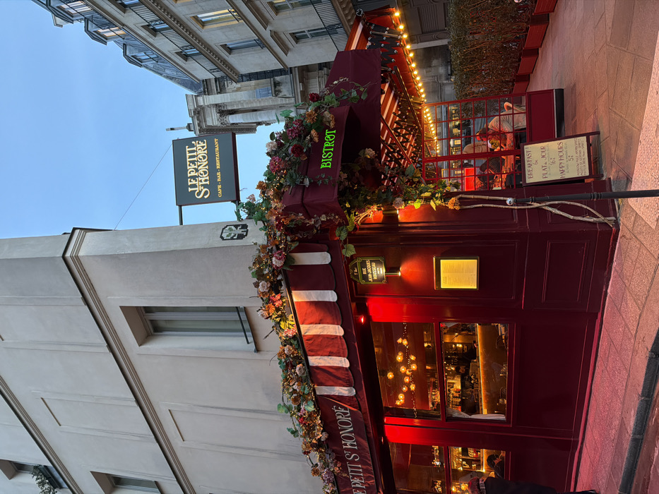The image showcases Le Petit S'Honore, a café-bar-restaurant with a bistro setup. The establishment features a red awning adorned with flowers and greenery. Signage indicates breakfast options and happy hour timings. The building has a vintage charm, and the interior is illuminated with warm lighting. There's also signage with text stating "E Petit S'Honore" and "Rue Jacques Rousseau."