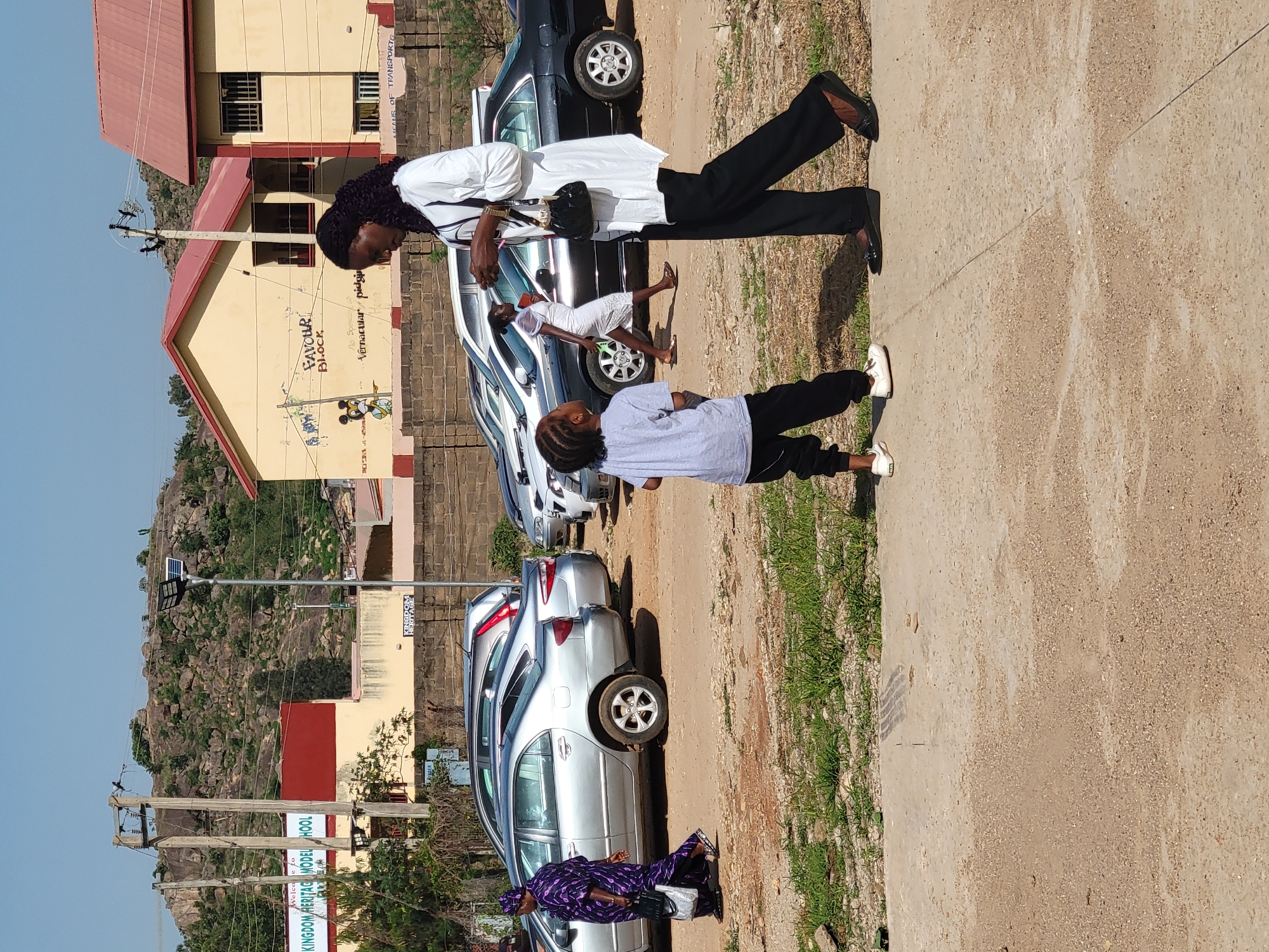 The image depicts a street scene in Gura Top, Nigeria, alongside a row of parked cars. A child stands facing a woman wearing a white top and black pants, who appears to be looking down at them. Another individual, adorned in a patterned purple outfit, is visible near the front of the parked vehicles. In the background, there's a building with the words "FAVOUR BLOCK" painted on it, along with an image of a cartoon character. A rocky hill is visible further in the background. Power lines and a sign reading "KINGDOM HERITAGE MODEL SCHOOL" are also present. The street is paved with a concrete sidewalk, with some grass growing along the edge.