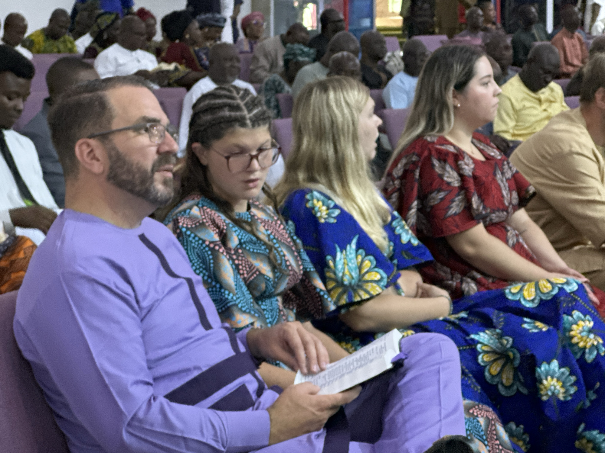 In an indoor setting, likely a place of worship or community gathering, a group of people are seated. The foreground is dominated by a man wearing a purple long-sleeved shirt with dark stripes. He has a beard and glasses and is holding a book open in his lap. To his right, a young woman with braided hair and glasses also appears to be looking at something in her lap, wearing a colorful patterned top. Further to the right, a woman with blonde hair is seen in profile, wearing a red patterned dress. Behind them, a crowd of individuals are visible, some in traditional attire, with varying skin tones. The seating appears to be upholstered in a muted color. The lighting is moderate, suggesting an indoor event.