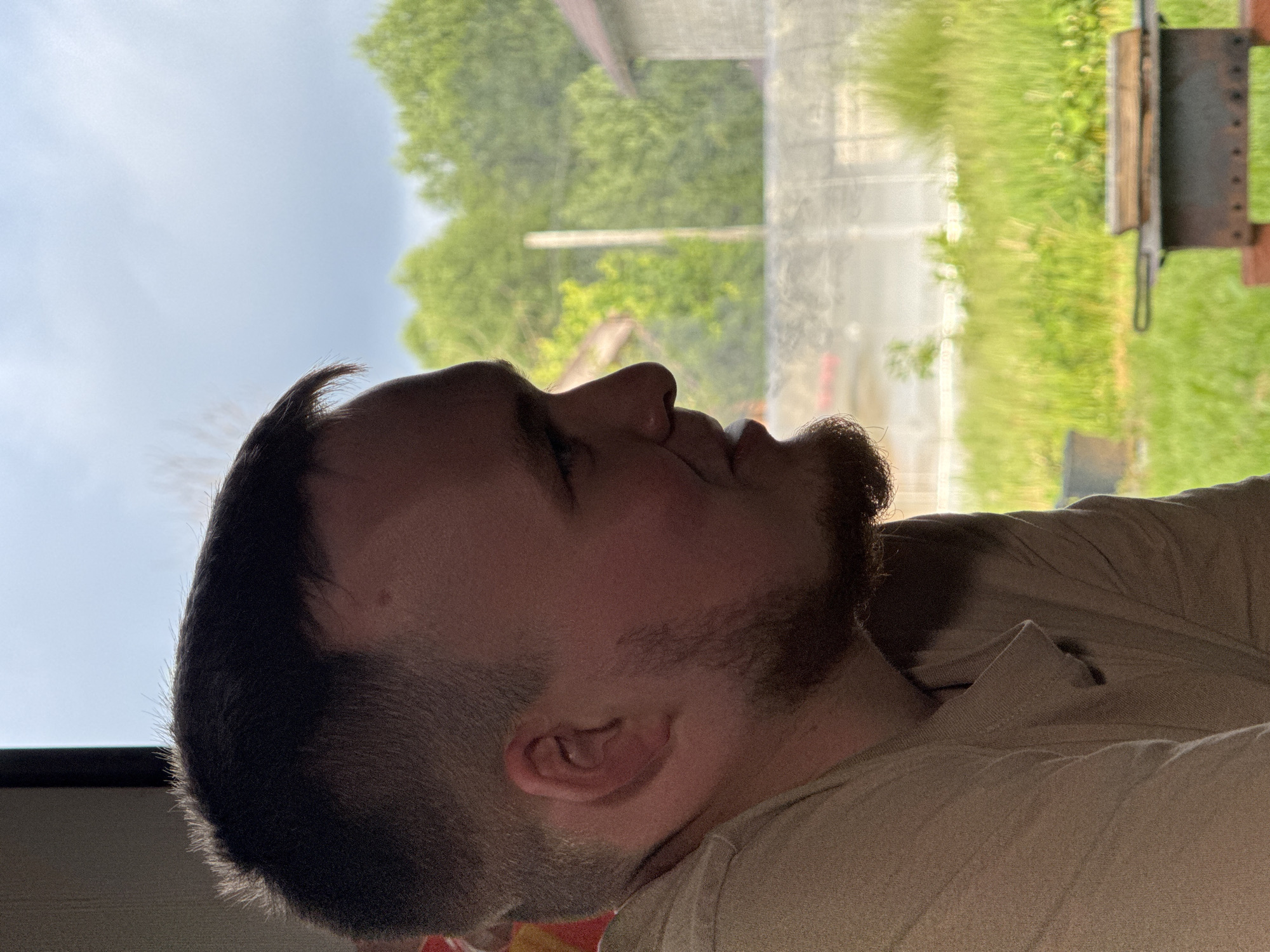 The image depicts a male individual in profile, facing right, looking out of a window in Krasnokamsk, Russia. The individual has short, dark hair with a faded cut and a trimmed beard. A distinct tan line is visible across his forehead, indicative of recent sun exposure. He wears a light-colored, possibly beige or tan, short-sleeved collared shirt. His expression is neutral to slightly smiling, with his mouth slightly ajar.
Through the window, an outdoor environment is visible under an overcast, light blue-grey sky. The landscape is predominantly green, featuring lush trees and ground covered in grass and some taller vegetation. In the mid-ground, a dark metal mangal (grill) with wooden pieces on its surface is positioned on a reddish base. Behind the mangal, a blurry grey concrete or stone structure, possibly a wall or fence, is present. Further in the background, a light-colored vertical pole is visible, partially obscured by foliage. To the upper right, a portion of a building with a reddish-brown roof and grey block walls is discernible. A dark, indistinct container, possibly a pot or bucket, is visible on the ground outside the window to the lower left.