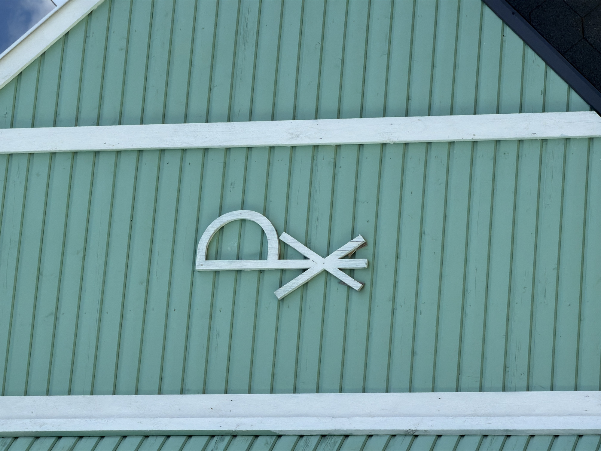 A close-up, eye-level exterior shot shows a light blue-green wooden clapboard wall of a building. A white wooden Chi Rho symbol is affixed to the wall, off-center to the right. The symbol is composed of a Greek chi (Χ) superimposed on a Greek rho (Ρ). The left edge of the building features a vertical white wooden trim. A dark, shingled roofline is visible in the lower right corner of the frame, intersecting the wall at an angle. The image is taken on a bright, possibly sunny day, with a patch of sky visible in the upper right corner. The location is the Church of the Assumption of the Most Holy Mother of God in Läskelä, Russia.