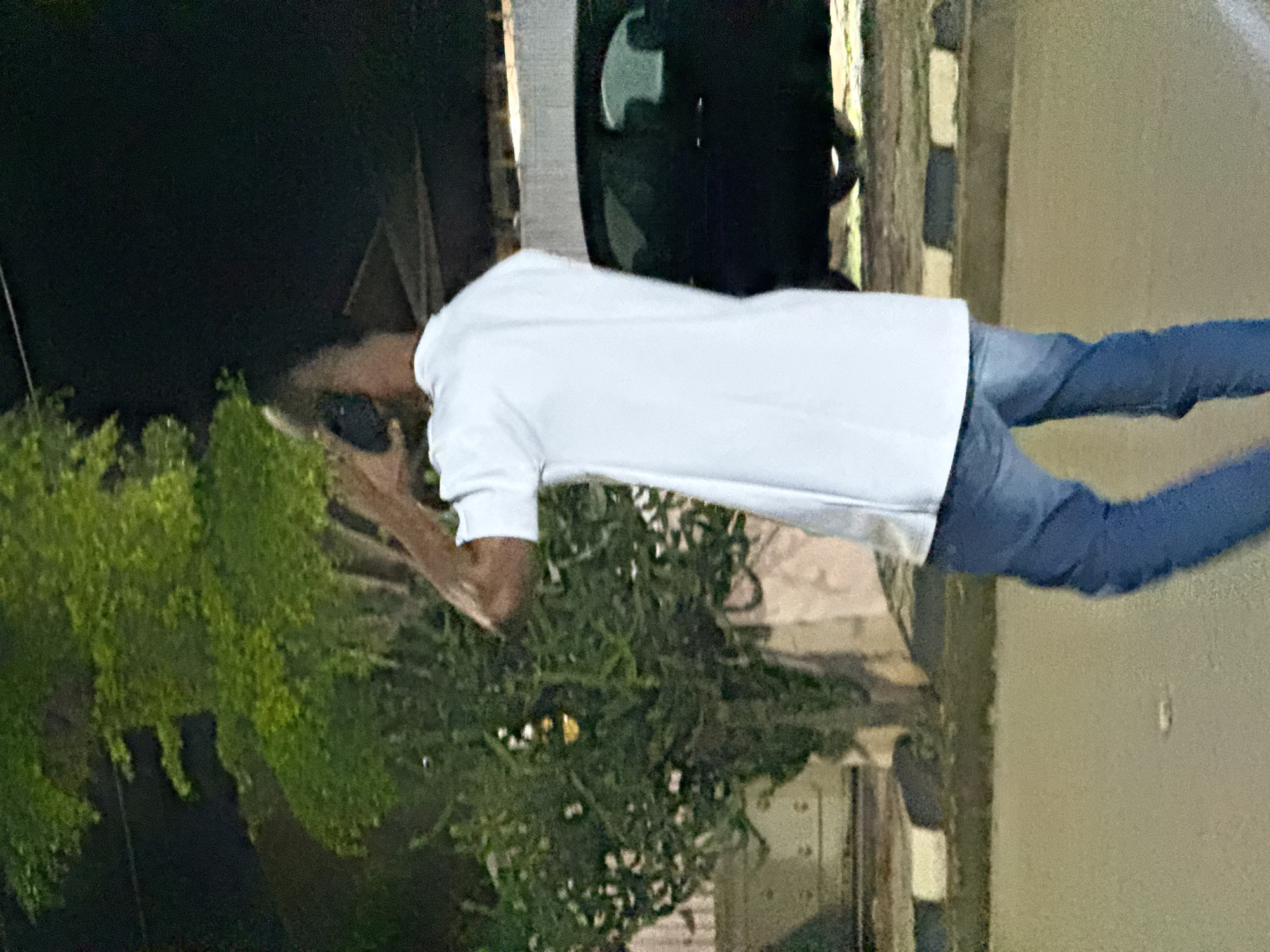 A person is standing outdoors at night, facing away from the camera, holding a phone to their ear. They are wearing a white t-shirt and blue jeans. Behind them, there is a dark-colored vehicle and a leafy green tree with some lights visible within its branches. The ground appears to be paved, with a curb in the background. The scene suggests an urban or suburban setting, likely in Abuja, Nigeria, given the context.
