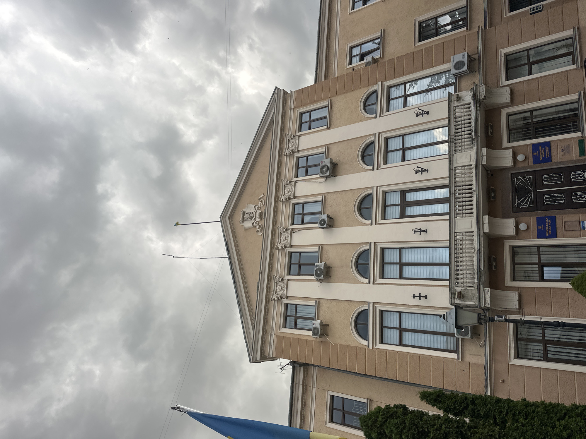 The image shows the Ternopil City Council building, located in Ternopil, Ukraine. It is a two-story building with a beige facade, white trim, and arched windows on the upper floor. A balcony with a decorative railing spans the front of the building. The sky is overcast and cloudy. A Ukrainian flag is visible on the left side of the image, partially obscuring the building. There are several air conditioning units mounted on the exterior walls. A street lamp stands in front of the building. Signs with Ukrainian text are visible on the facade.