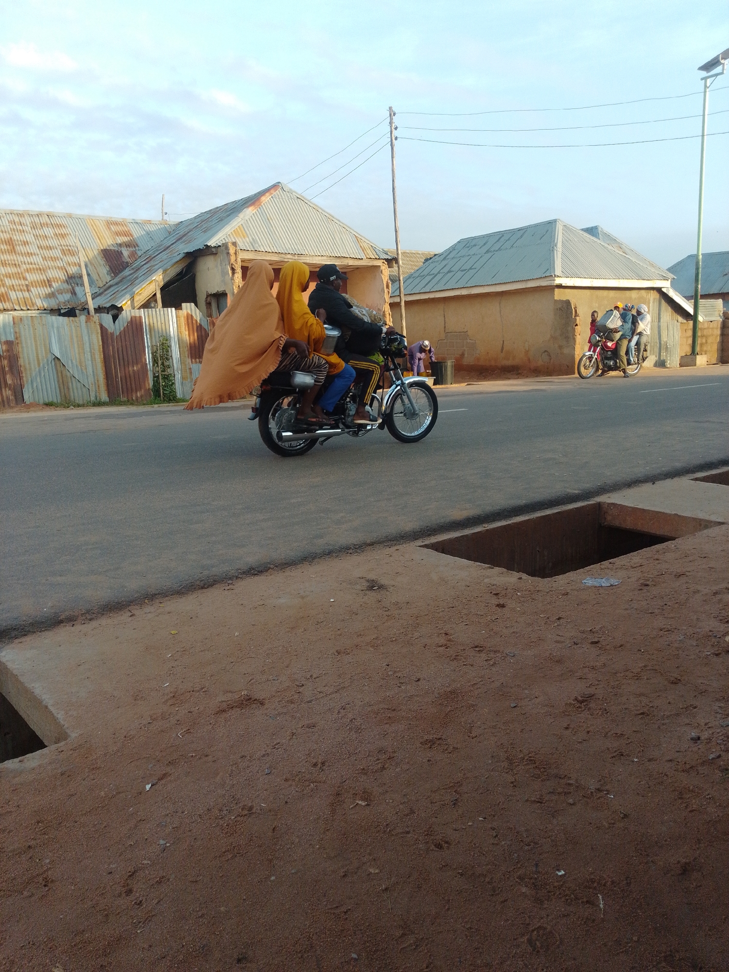 The image depicts a street scene in Tempure, Nigeria. In the foreground, there is a dirt ground with a large rectangular hole, possibly a drain or construction site. The street is paved with asphalt. On the street, there are several motorcycles and people.
The main focus is on a motorcycle carrying three individuals. The driver, wearing a dark helmet, is at the front. Behind him, a person in a bright orange hijab and a yellow garment is seated. Another person, partially obscured, is seated behind the person in the hijab. The motorcycle is red and appears to be in motion.
To the right of this motorcycle, another red motorcycle is parked, with two people seated on it, both wearing helmets. Further down the street, a third motorcycle, a lighter color, is visible with a single rider.
The background consists of buildings with corrugated metal roofs and walls made of mud bricks. The sky is bright with some clouds. There are utility poles with wires running across the scene. The overall impression is one of daily life and transportation in a Nigerian town.