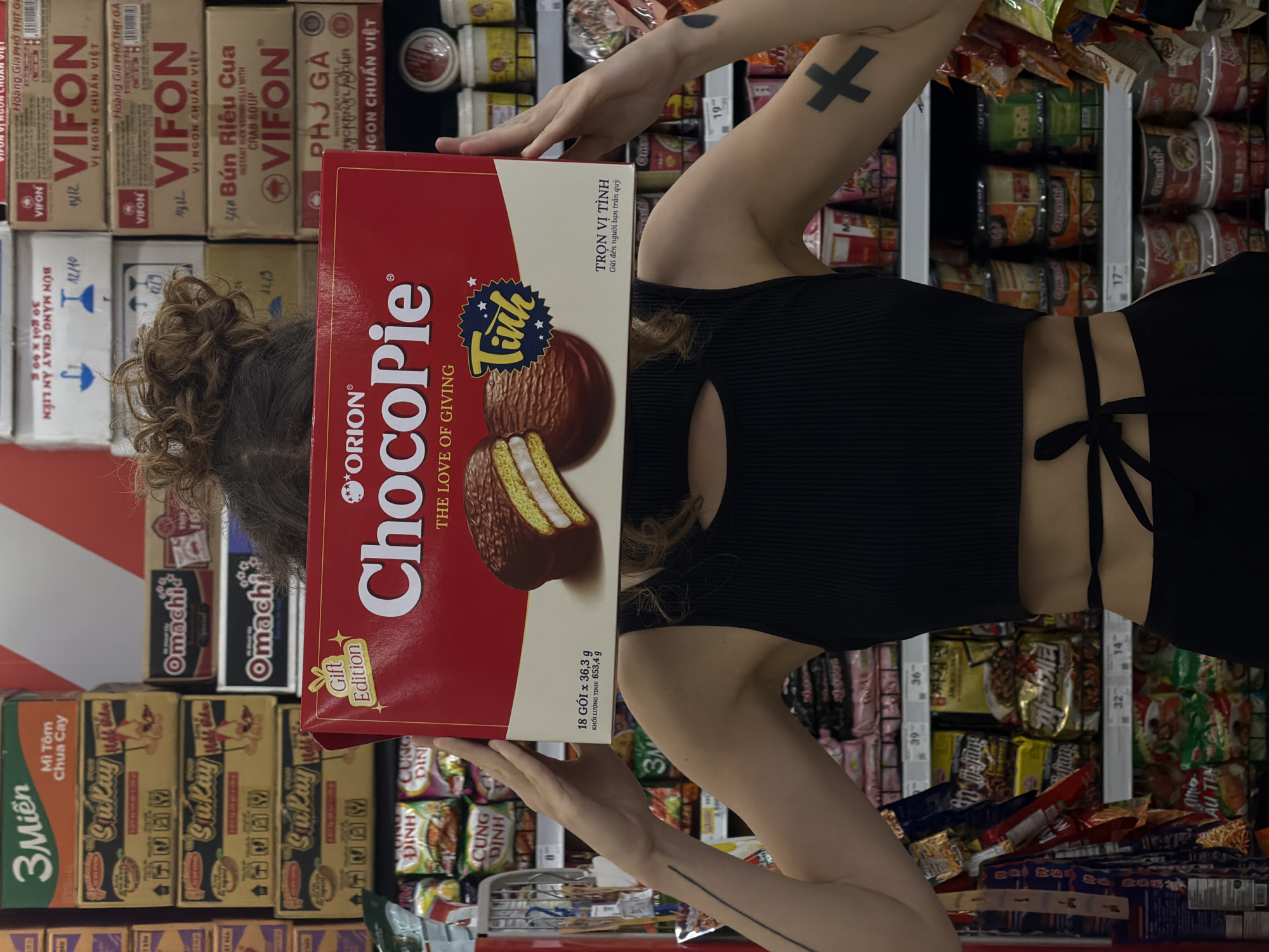 An image, rotated 90 degrees counter-clockwise, depicts a person in a supermarket aisle in Đà Nẵng, Vietnam, holding a large red box of Orion ChocoPie. The person's face is obscured by the box, which is held up, likely for examination. The subject wears a black ribbed top with tie details at the back, and their dark, curly hair is pulled back in a bun. Visible tattoos include a black 'X' on their upper right arm and a thin black line extending down their left forearm.
The ChocoPie box is prominently displayed, featuring the brand name "ORION ChocoPie" and the slogan "THE LOVE OF GIVING," along with "Gift Edition." It shows an image of two ChocoPies, one whole and one cut to reveal the white cream filling. Vietnamese text on the box includes "Tinh," "TRỌN VỊ TÌNH," "Gửi đến người bạn trân quý," and "18 GÓI x 36,3 g," indicating a total net weight of "KHỐI LƯỢNG TỊNH: 653,4 g."
The background reveals typical supermarket shelves stocked with numerous packaged food items, predominantly instant noodles and dry goods. Brands visible include VIFON (with varieties like "Hoàng Gia PHỞ THỊT GÀ," "Hoàng Gia PHỞ THỞ BÒ," "Bún Riêu Cua," and "PHỞ GÀ"), Omachi, 3 Miền ("Mì Tôm Chua Cay"), SuKay, CUNG DINH, and JJA PAGHETTI (짜파게티). Price tags are visible on the shelves, showing numbers like "17-," "19-," "32-," "36-," and "39-." The lighting is bright and even, consistent with an indoor retail environment.