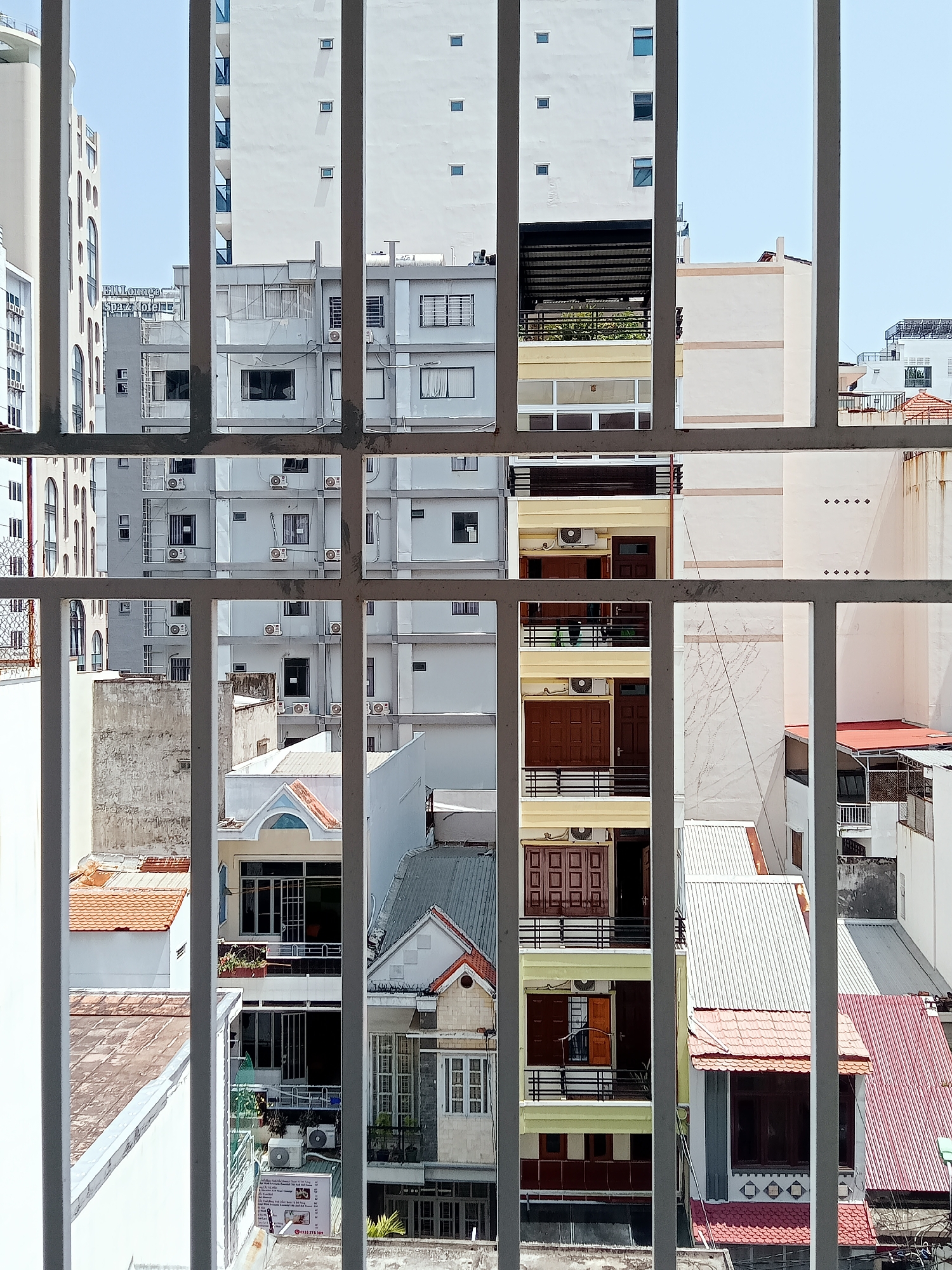 The image depicts a view of a densely packed urban landscape in Nha Trang, Vietnam, as seen through a metal security grate. The foreground shows the grate itself, composed of vertical bars and horizontal cross pieces. Beyond the grate, the scene reveals a jumble of buildings of varying heights, styles, and materials. Some are modern high-rise apartments, primarily white or gray, with numerous windows and air conditioning units. Others are low-rise residential buildings, with tiled roofs and balconies. Colors range from muted grays and whites to splashes of yellow and red from the smaller buildings.
The buildings are closely spaced, with little visible greenery. The sky is light blue and relatively clear. The overall impression is one of a crowded, bustling urban environment typical of many Southeast Asian cities. There are no people visible in the frame.