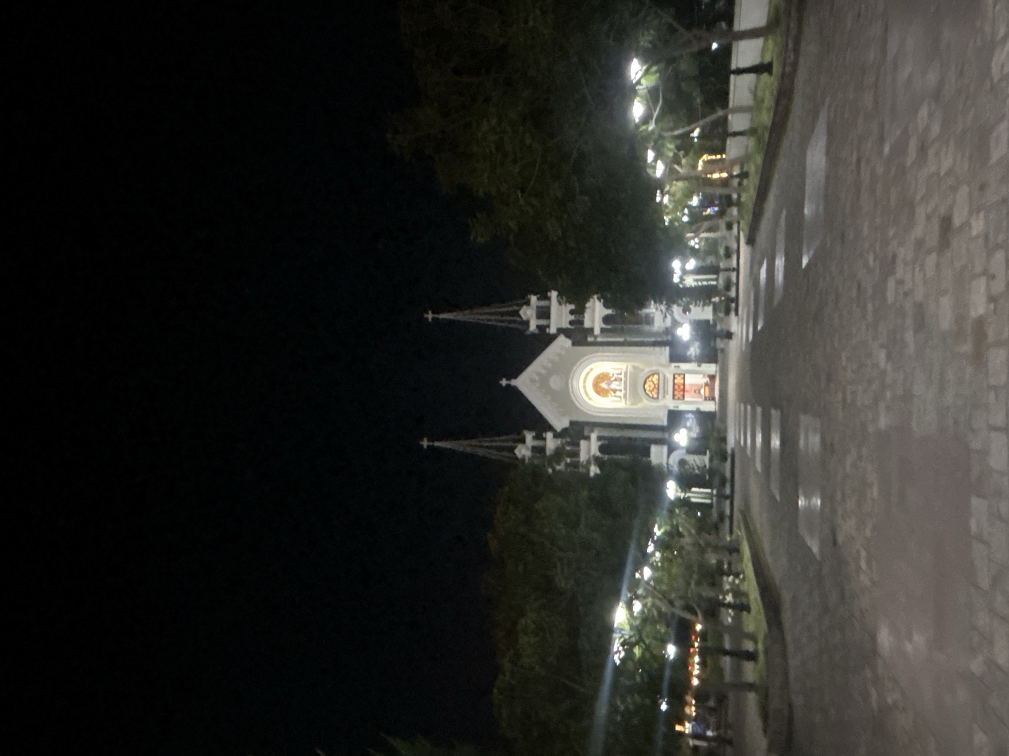 The image depicts a church at night in the Bac Natural Heritage Area Trang, Vietnam. The church is brightly lit, with two prominent spires. Trees and lampposts line the walkway leading to the church. The foreground shows a paved area. The surrounding environment is dark.
