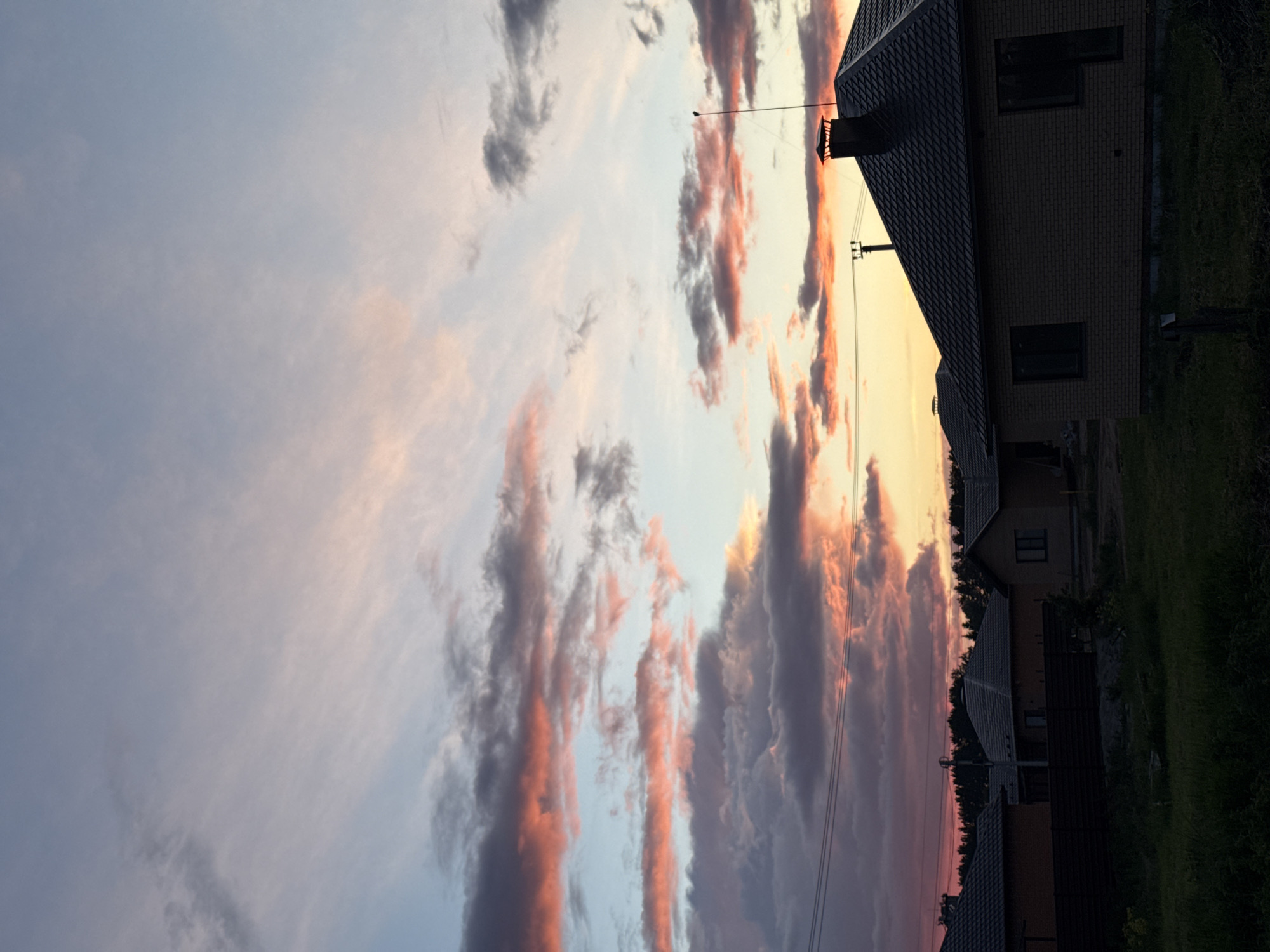 The image shows a residential area in what appears to be the city of Chaikovsky, Russia, judging by the local architectural style. The sky is filled with clouds at sunset, with hues of orange and pink near the horizon. Several houses are visible along the right side, some with dark roofs and windows. Grass and trees flank the houses. Power lines stretch across the sky near the houses.