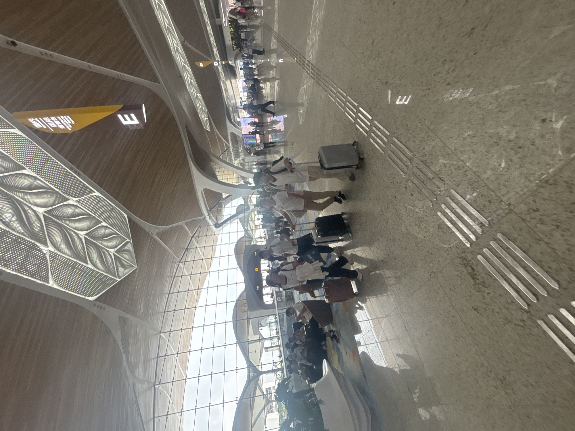 The image depicts the interior of a modern airport terminal, likely located in Baiyun District, China, with a high, arched ceiling constructed from wood and metal. Natural light streams in through a large, curved glass facade, illuminating the polished concrete floor. Several individuals are visible, some walking with luggage, others seated in lounge areas. Prominent signage hanging from the ceiling indicates gates and directions, including one with the letter "E" and Chinese characters. Tactile paving, designed for the visually impaired, is present on the floor. The overall atmosphere is one of bustling transit within a spacious, contemporary public building.