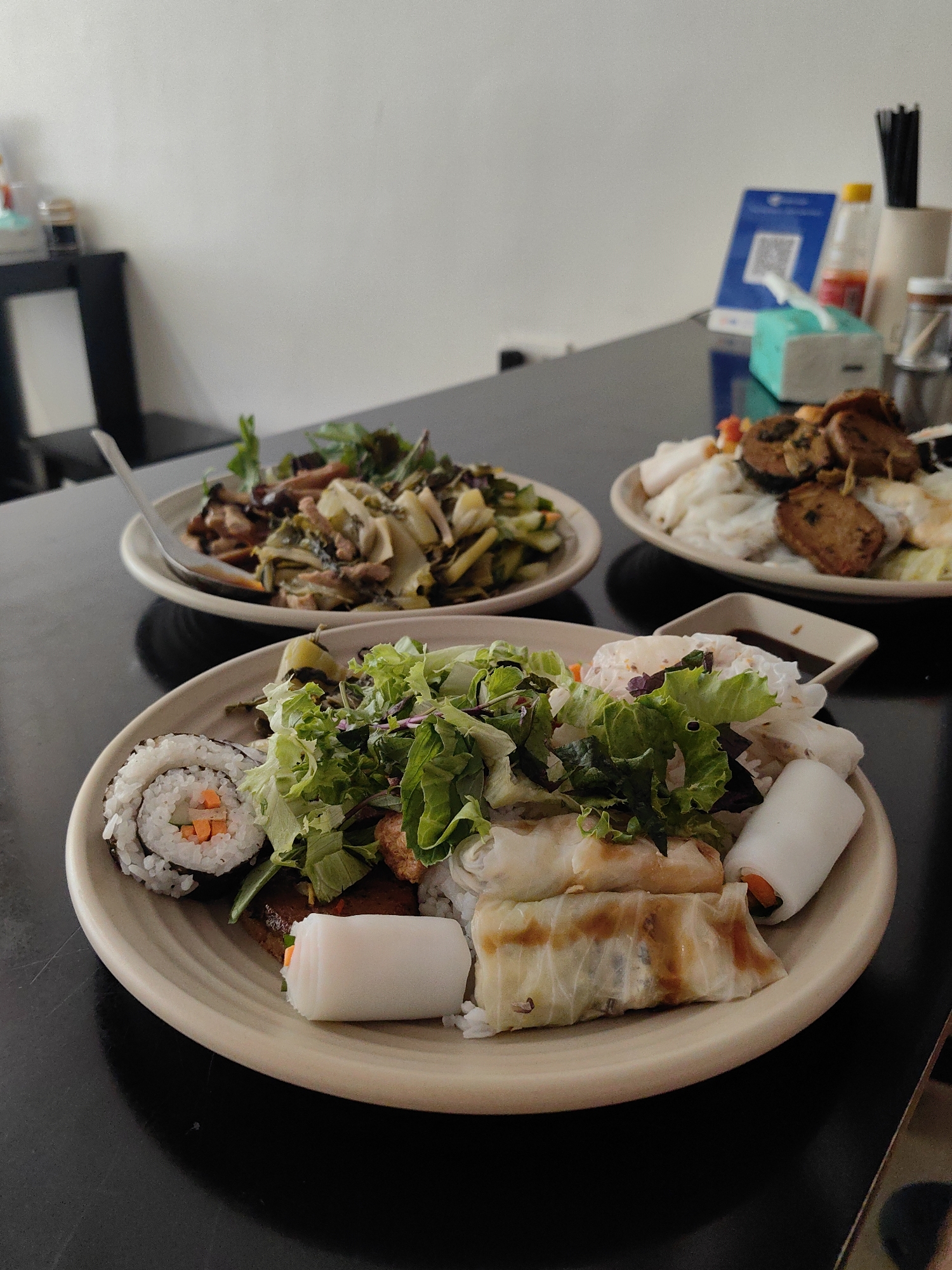 A table in Hong Gai Ward, Vietnam, holds three plates of food. The nearest plate contains sushi, leafy greens, and spring rolls. Another plate features a mixture of leafy greens and sauteed vegetables, with a fork. The furthest plate is filled with rice noodles, fried tofu, and leafy greens. A small dish of dipping sauce accompanies the food. Behind the plates are a blue QR code stand, a bottle of hot sauce, a tissue box, and other miscellaneous objects. The table has a glossy black surface.