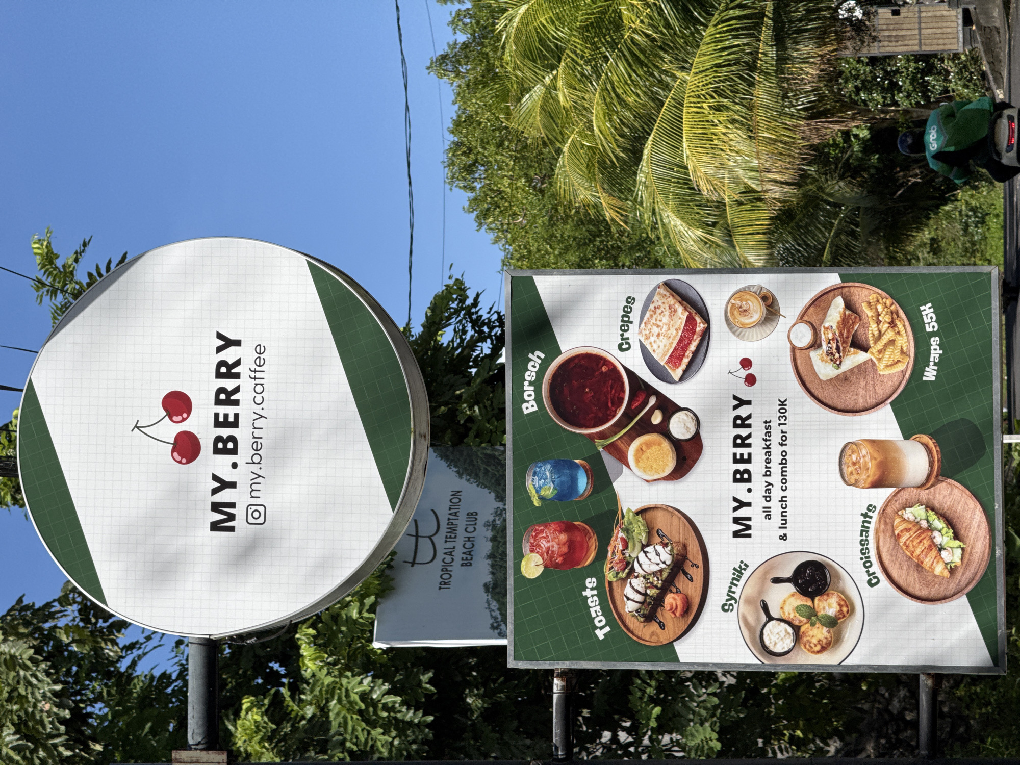 This outdoor, vertically-oriented image captures two prominent signs advertising businesses, set against a backdrop of lush tropical greenery and a clear blue sky.
The most prominent sign is a circular, white and dark green placard for "MY.BERRY," featuring a logo of two cherries and the text "MY.BERRY" with the Instagram handle "@my.berry.caffee". The sign has a subtle grid pattern on its white surface.
Adjacent to it, and partially obscuring another sign, is a large rectangular menu board, also for "MY.BERRY." This board displays various food and drink items with accompanying images and prices. Visible text includes "MY.BERRY all day breakfast & lunch combo for 130K," and specific items like "Borsch," "Crepes," "Wraps 55K," "Croissants," "Syrniki," and "Toasts." The images depict dishes such as borscht soup, colorful beverages, avocado toast, coffee, a crepe, a wrap with fries, an iced coffee, a croissant, and Syrniki pancakes.
Behind the circular MY.BERRY sign, a smaller white sign is partially visible, reading "TROPICAL TEMPTATION BEACH CLUB."
The background is dominated by vibrant green foliage, including large palm fronds and other trees, under a bright, cloudless blue sky. Overhead power lines traverse the sky. In the upper right corner, a person wearing a green jacket, likely a delivery driver, is seen on a scooter with the "Grab" logo visible, hinting at the presence of a delivery service.
The strong sunlight and clear sky suggest it is daytime, possibly mid-morning or afternoon. The tropical vegetation and text cues like "BEACH CLUB" and prices in "K" (indicating Indonesian Rupiah) point to a setting in a tropical region, consistent with Kuta Selatan, Indonesia.