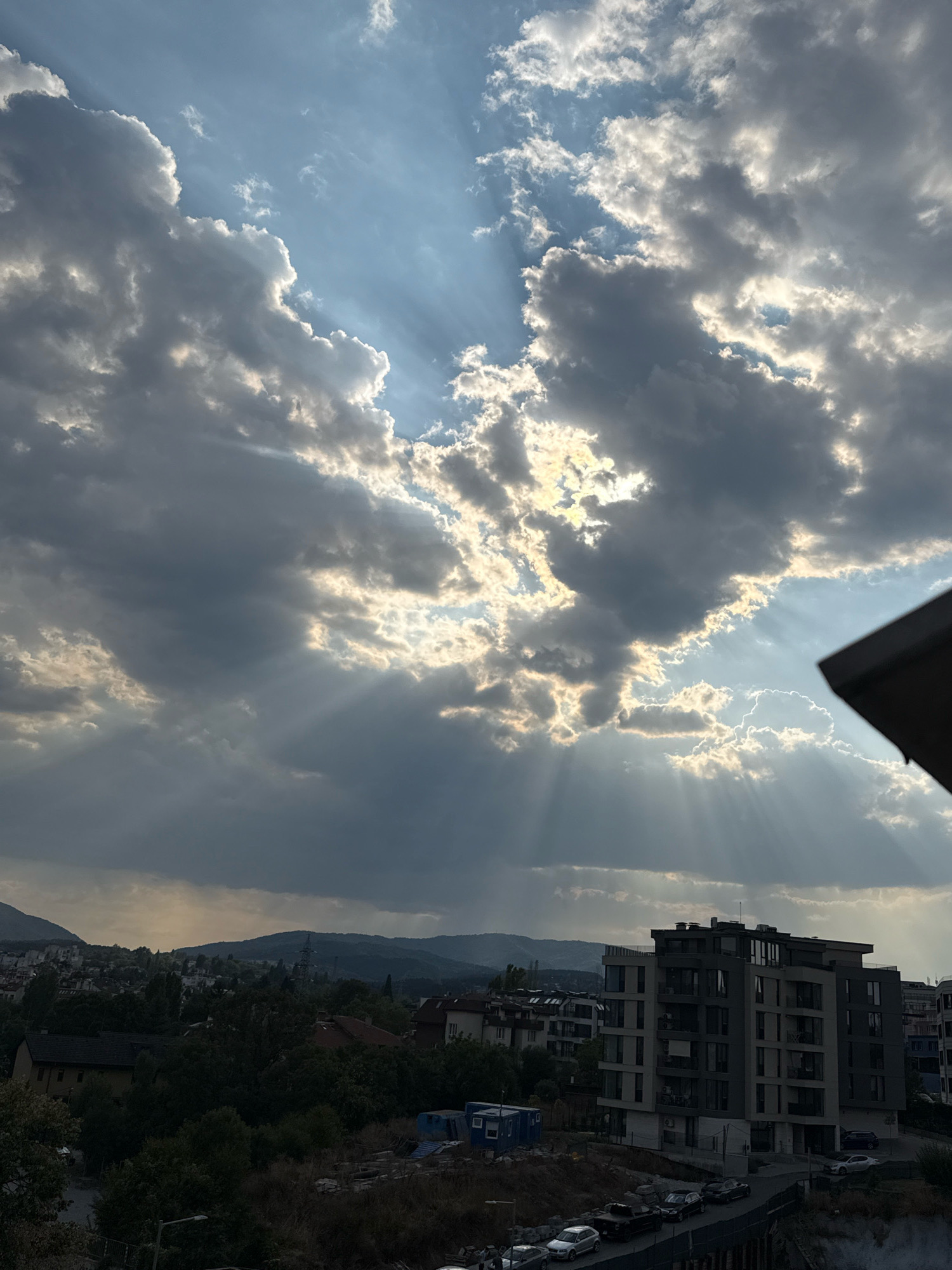 The image captures a dramatic sky over a city, likely Sofia, Bulgaria, characterized by crepuscular rays emanating from behind large, textured clouds. The sun, partially obscured, casts beams of light through openings in the cloud cover, creating a luminous effect that illuminates portions of the sky and casts shadows.
In the foreground, the roof of a building juts into the frame from the upper right corner. Below the sky, a cityscape unfolds. Buildings of varying heights and architectural styles are visible, some appearing to be modern apartment complexes with dark facades and numerous windows, while others are older structures with red-tiled roofs. Dense green trees populate the landscape between the buildings, interspersed with patches of drier vegetation and construction debris. A road or pathway winds through the scene, with several cars parked along its edge. In the distance, rolling hills or mountains form the backdrop, their shapes softened by the atmospheric conditions. The overall impression is a moment of transition, perhaps during sunrise or sunset, marked by the dynamic interplay of light and shadow in the sky and across the urban environment.