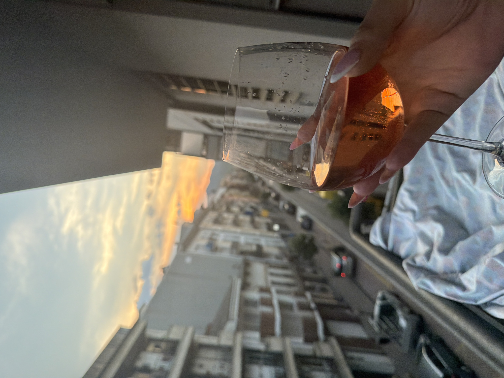 An extreme close-up captures a woman's hand, with long, light-pink manicured fingernails, holding a stemmed wine glass filled with a pinkish-orange liquid. The glass has condensation droplets on its exterior. The background is an out-of-focus urban scene, likely taken from an elevated position such as a balcony. Buildings line a street, with parked cars visible below. The sky above is a soft gradient of blue and orange, indicative of a sunset. The scene appears to be set in the city of Arbúcies, Spain, given the architectural style of the buildings and the general atmosphere.
